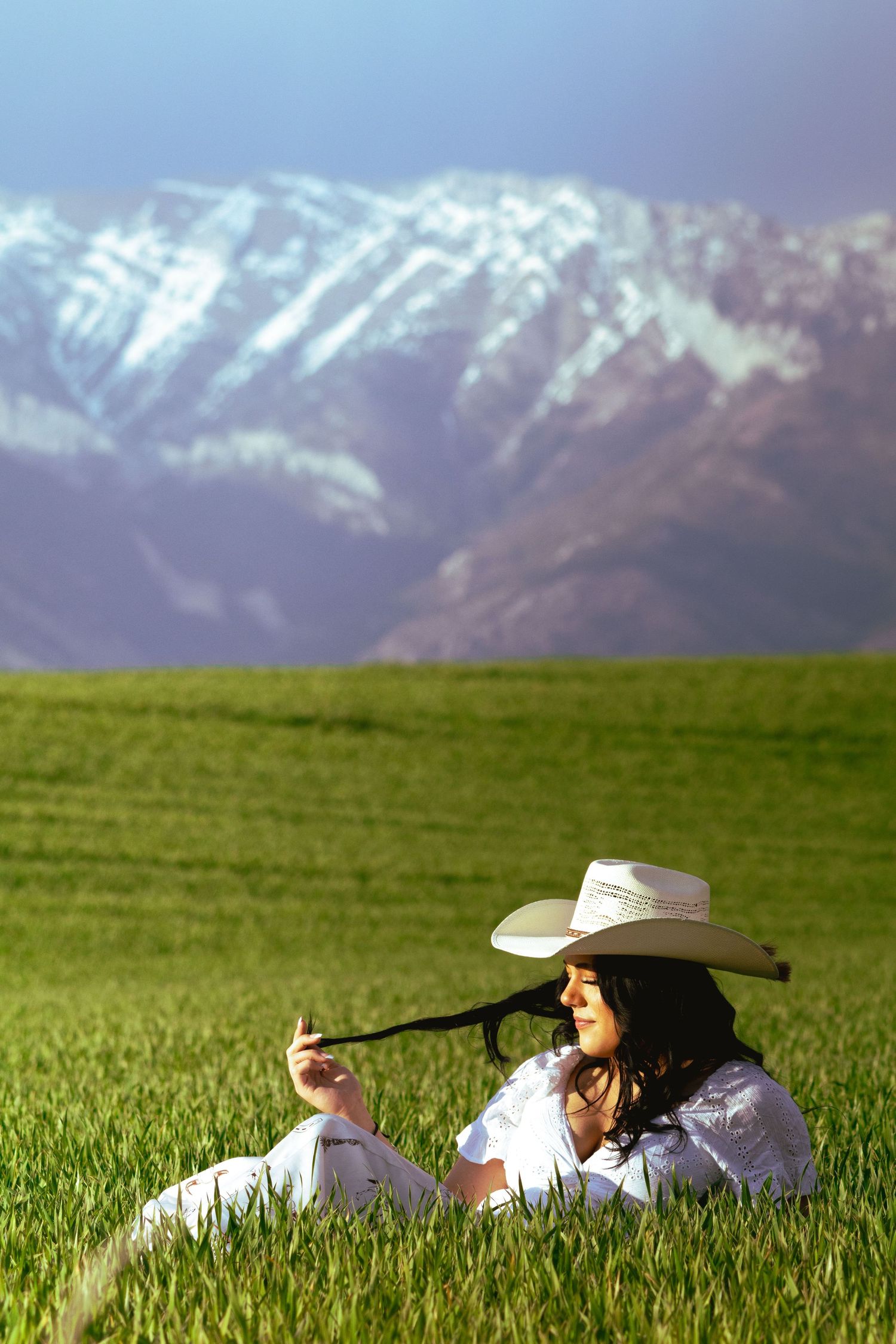 Young woman in white outfit and cowboy hat relaxing in a green field with snow-capped mountains behind her.