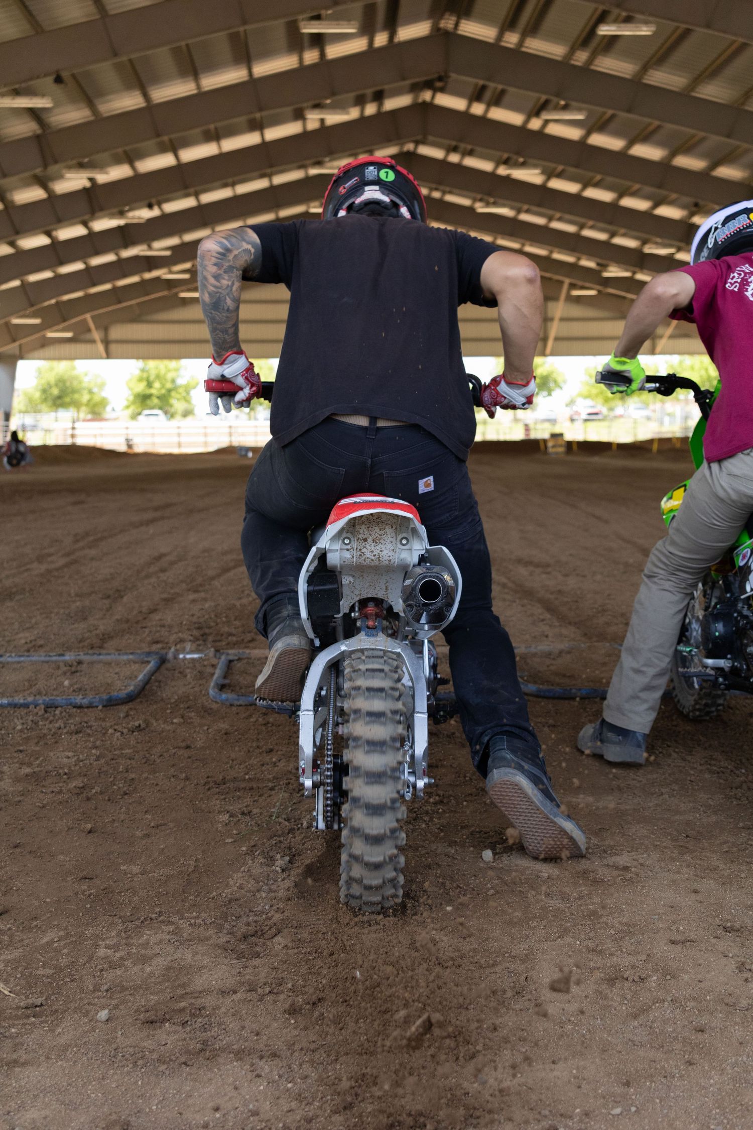 Rear view of motocross rider revving dirt bike engine at starting line in indoor arena.