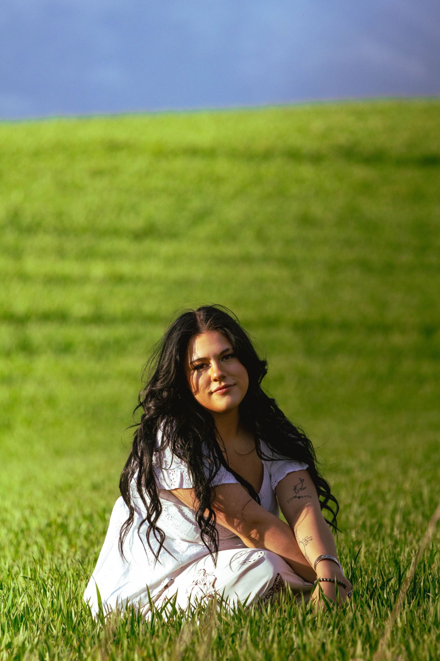 Young woman with long dark hair sitting cross-legged in a lush green field wearing white.