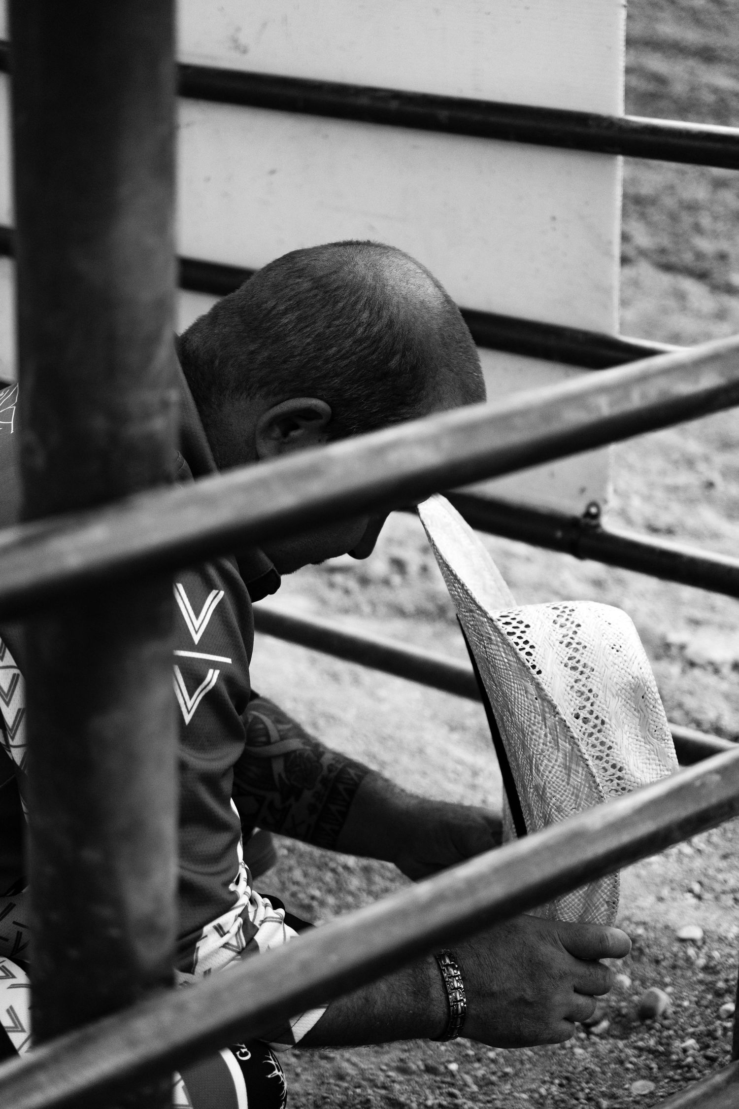A young child sits barefoot on the ground behind metal railings, holding a white hat in black and white photo.