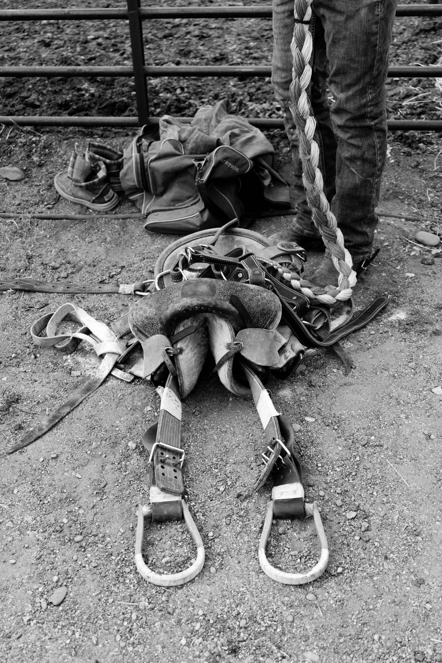 Worn rodeo riding gear including leather chaps and buckles lies on the ground beside a cowboys boots.