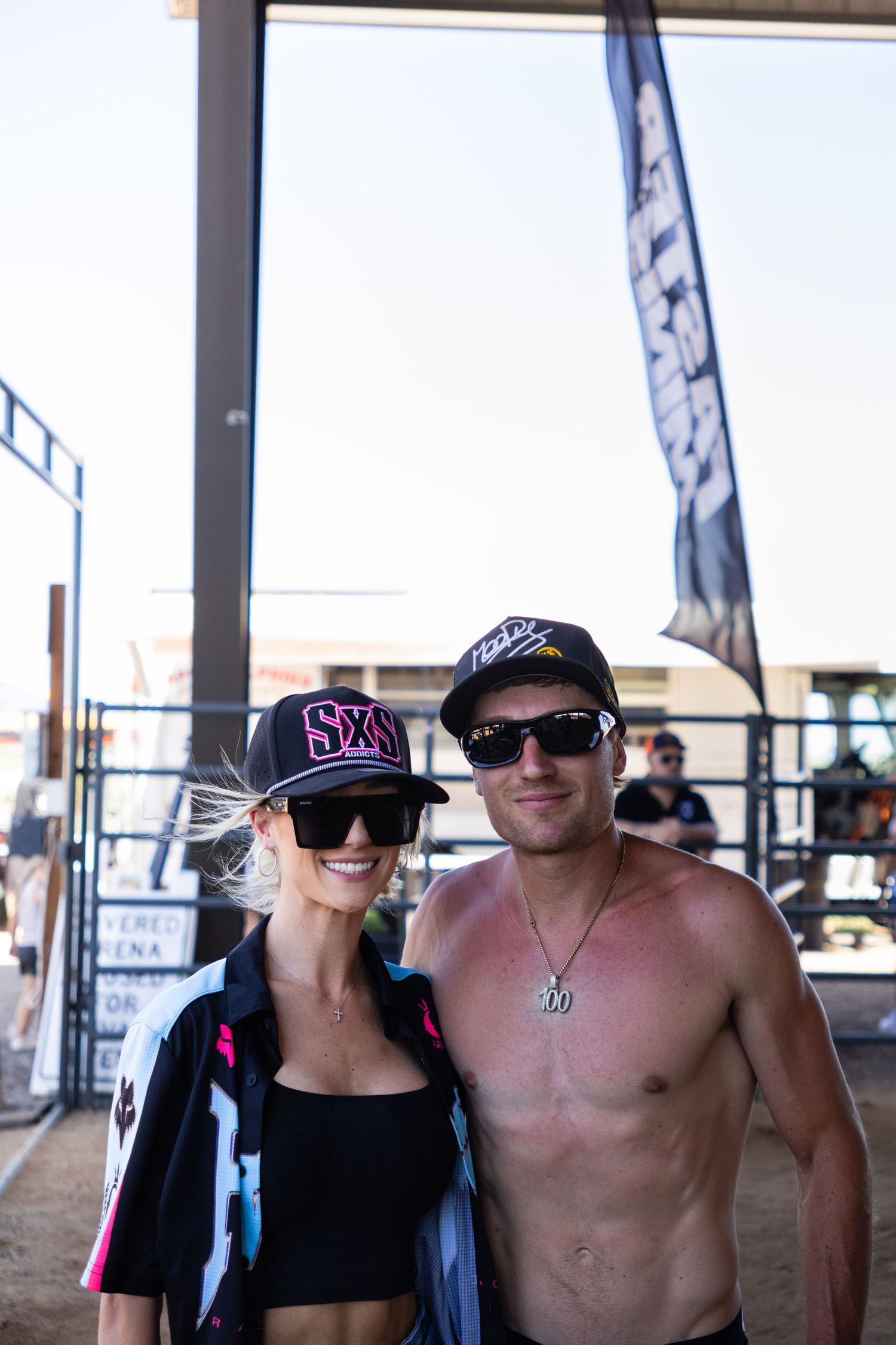 Smiling couple in snapback caps and sunglasses pose together at an outdoor motocross event.