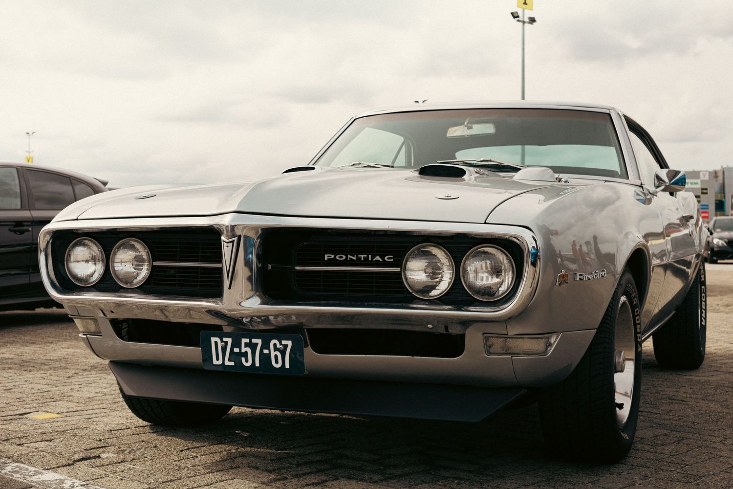 Vintage silver Pontiac Firebird muscle car parked on pavement with Dutch license plate DZ-57-G7.