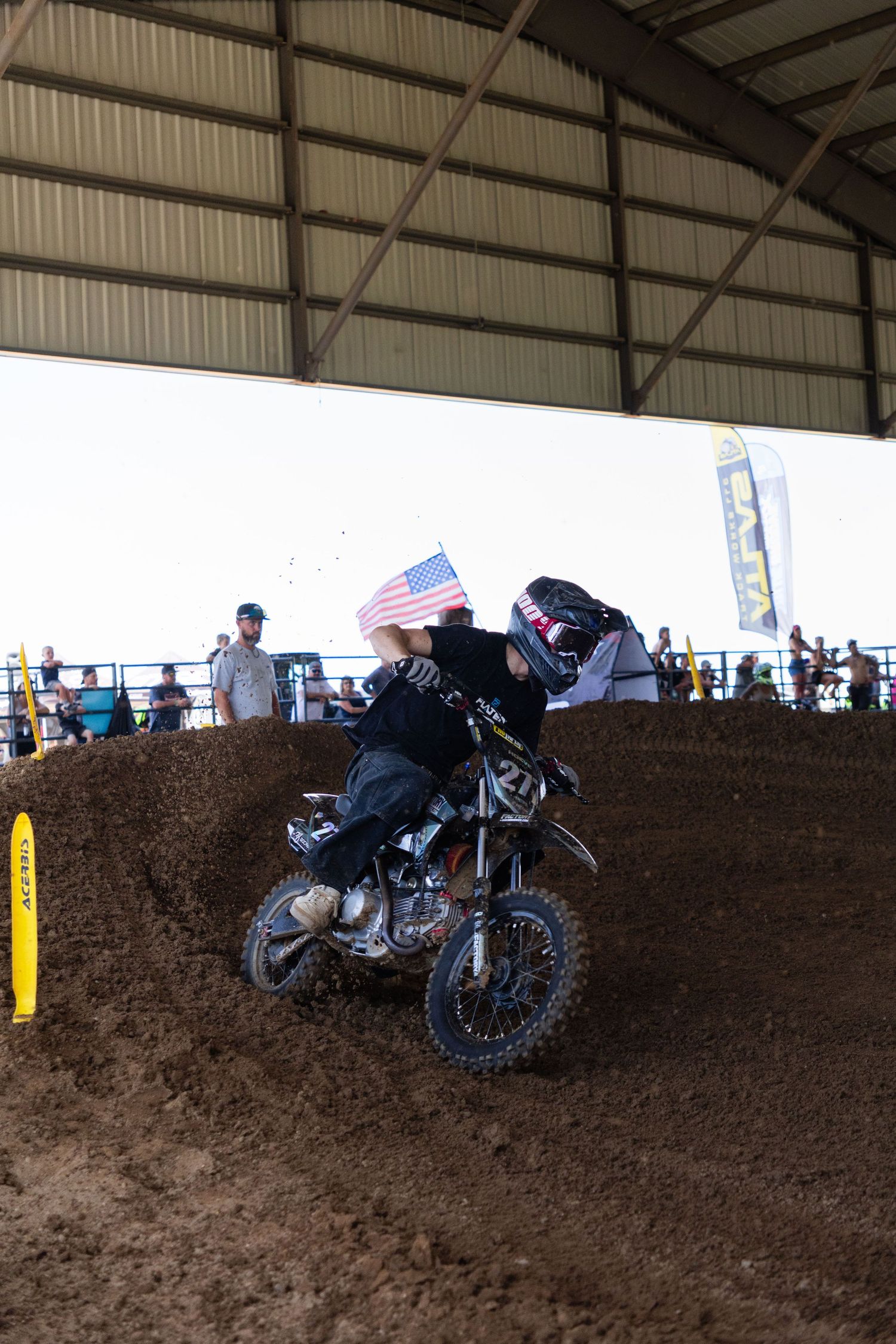 Motocross rider waves American flag while riding over dirt berm inside covered arena at competition.