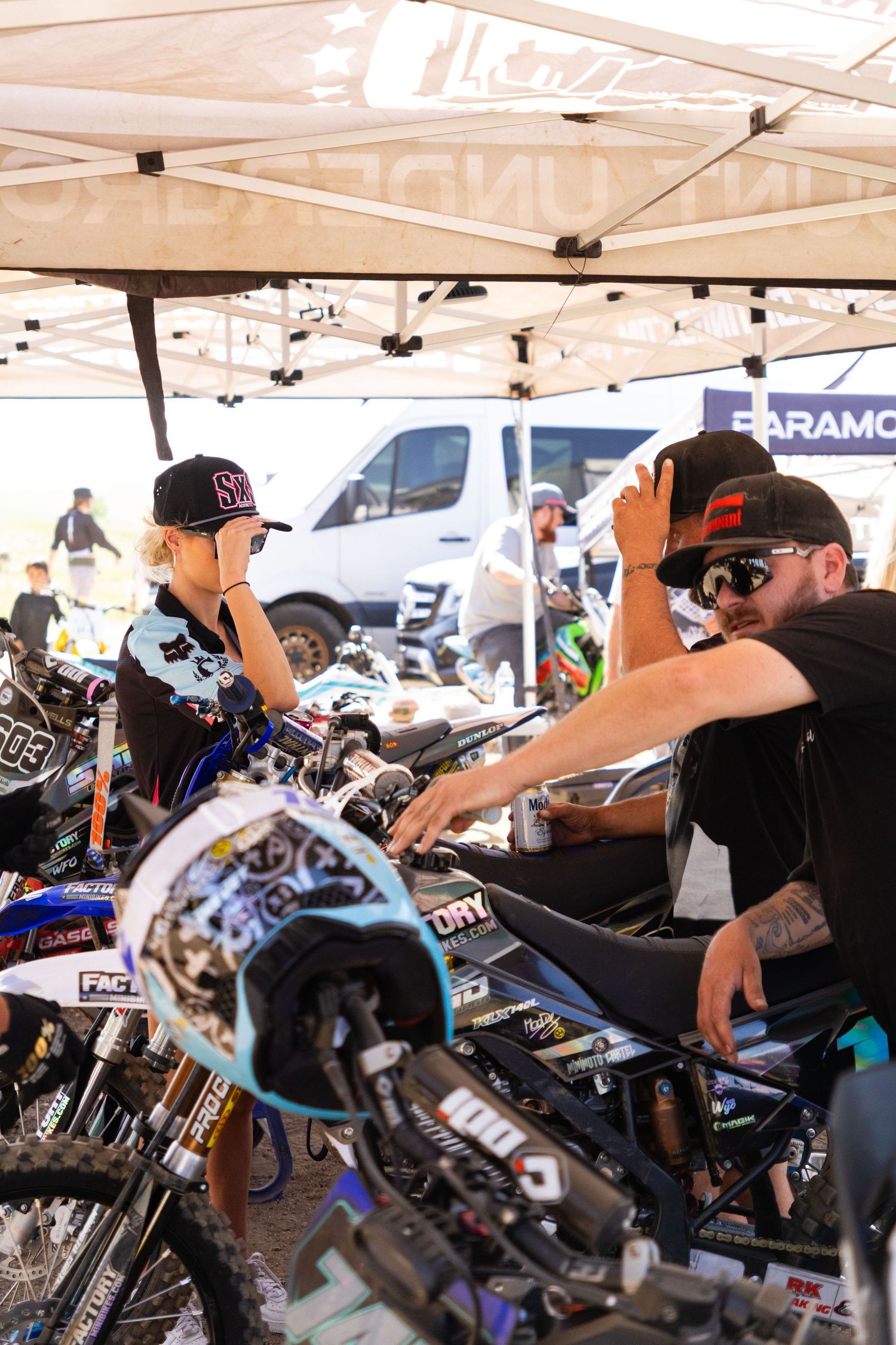 Two people inspecting dirt bikes at a motocross event under a canopy tent.