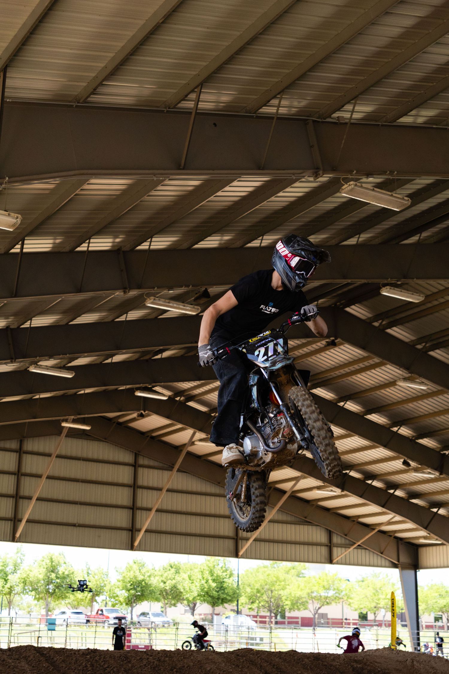 Motocross rider in black gear jumping dirt bike high inside covered indoor motocross facility.