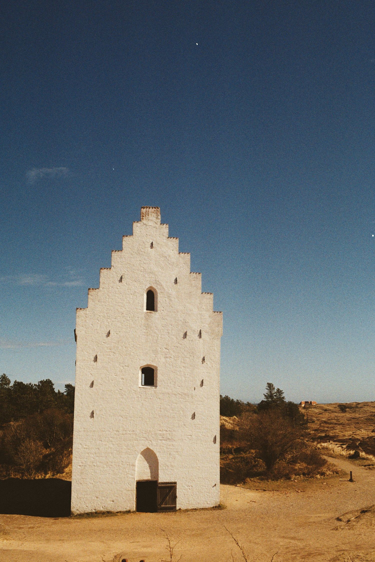 White Danish stepped gable church tower rising against clear blue sky in sandy landscape near Skagen, Denmark.
