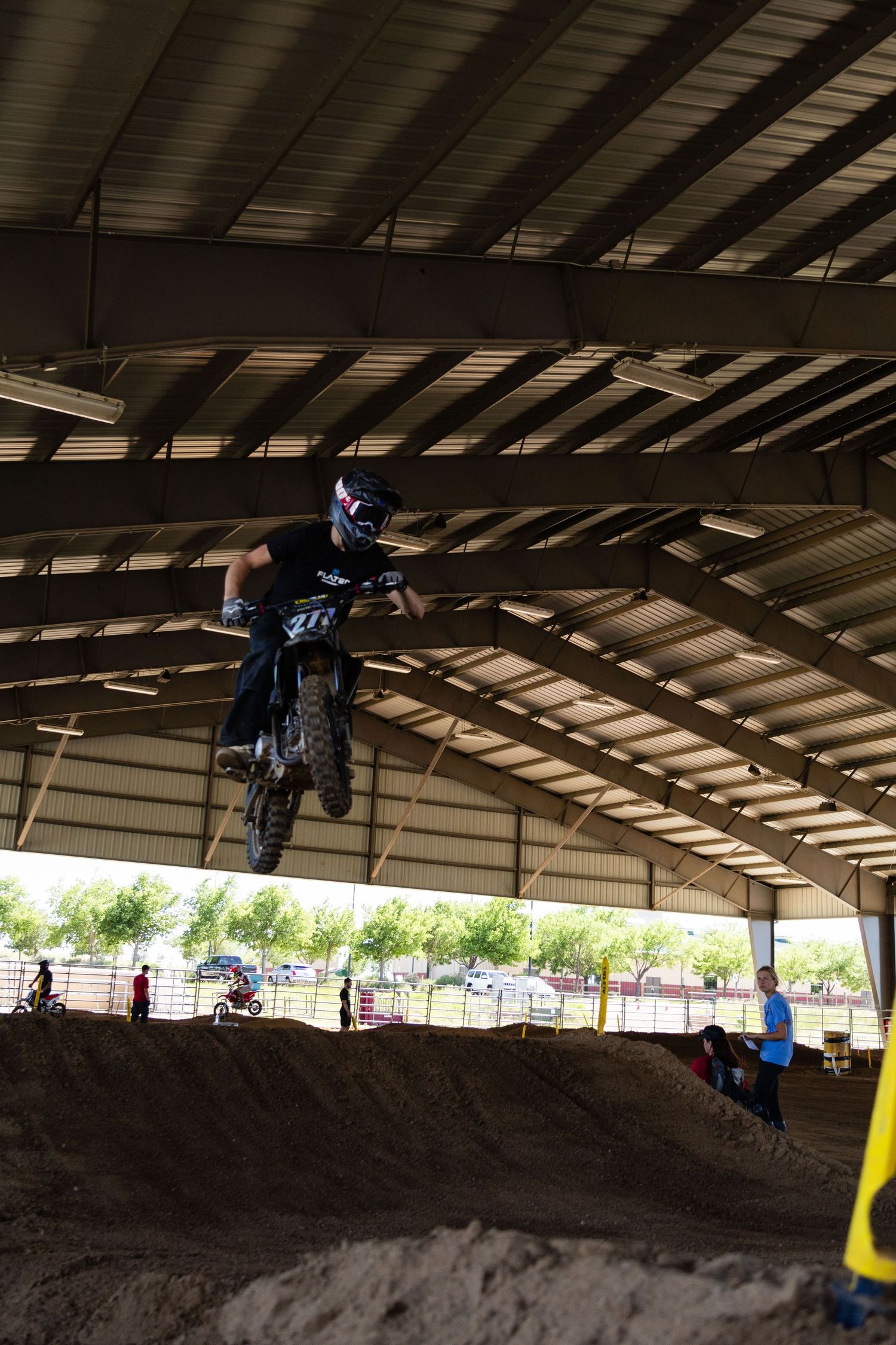 Motocross rider in black gear launches off dirt jump at indoor supercross arena.