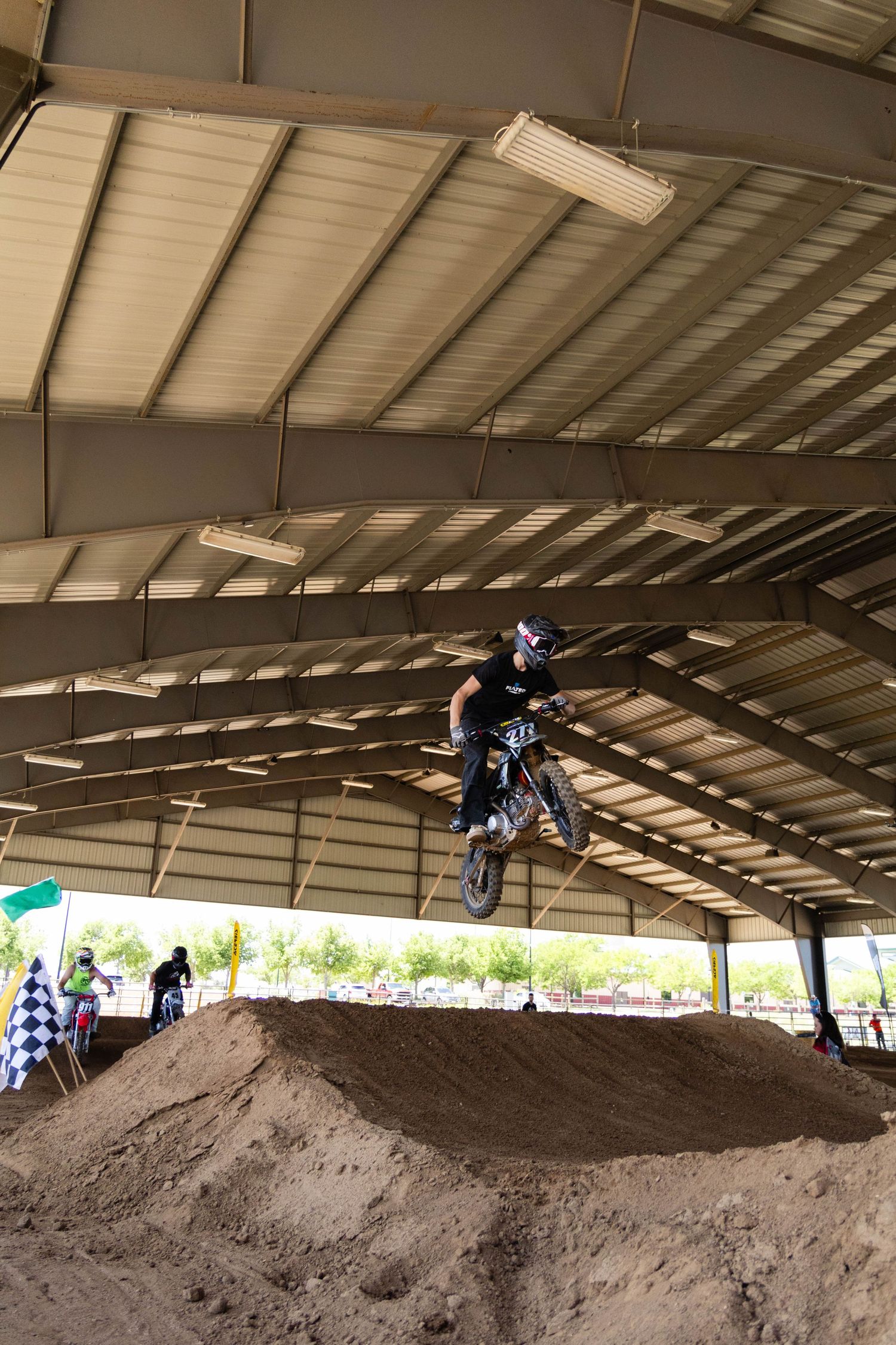 Motocross rider launching off dirt jump inside covered arena with checkered flag visible.