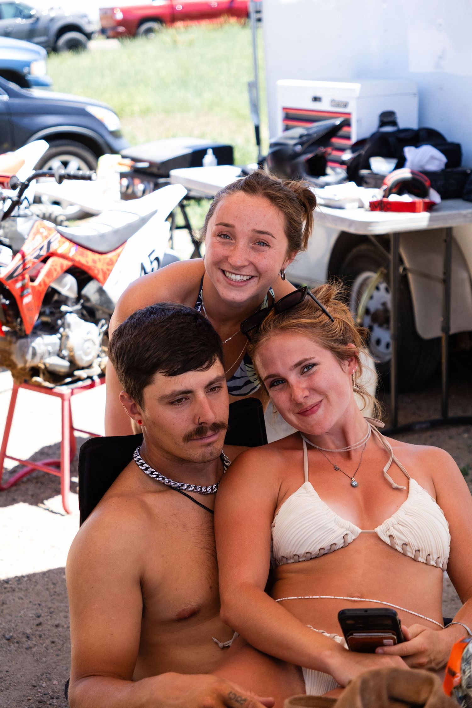 Three young people smiling together at a motocross racing event with dirt bikes in the background.