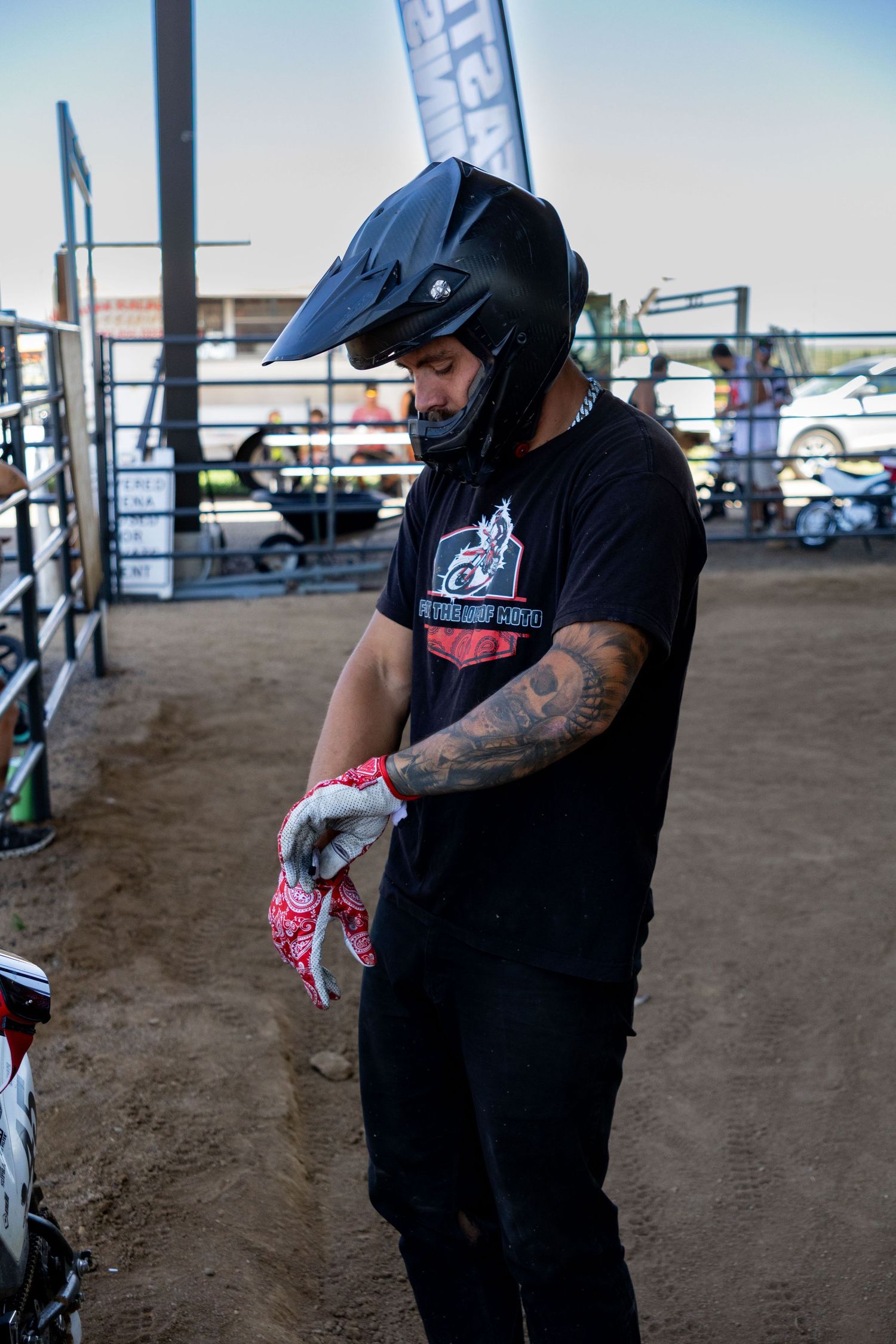 Tattooed motocross rider in black helmet and team shirt puts on gloves before race at dirt event.