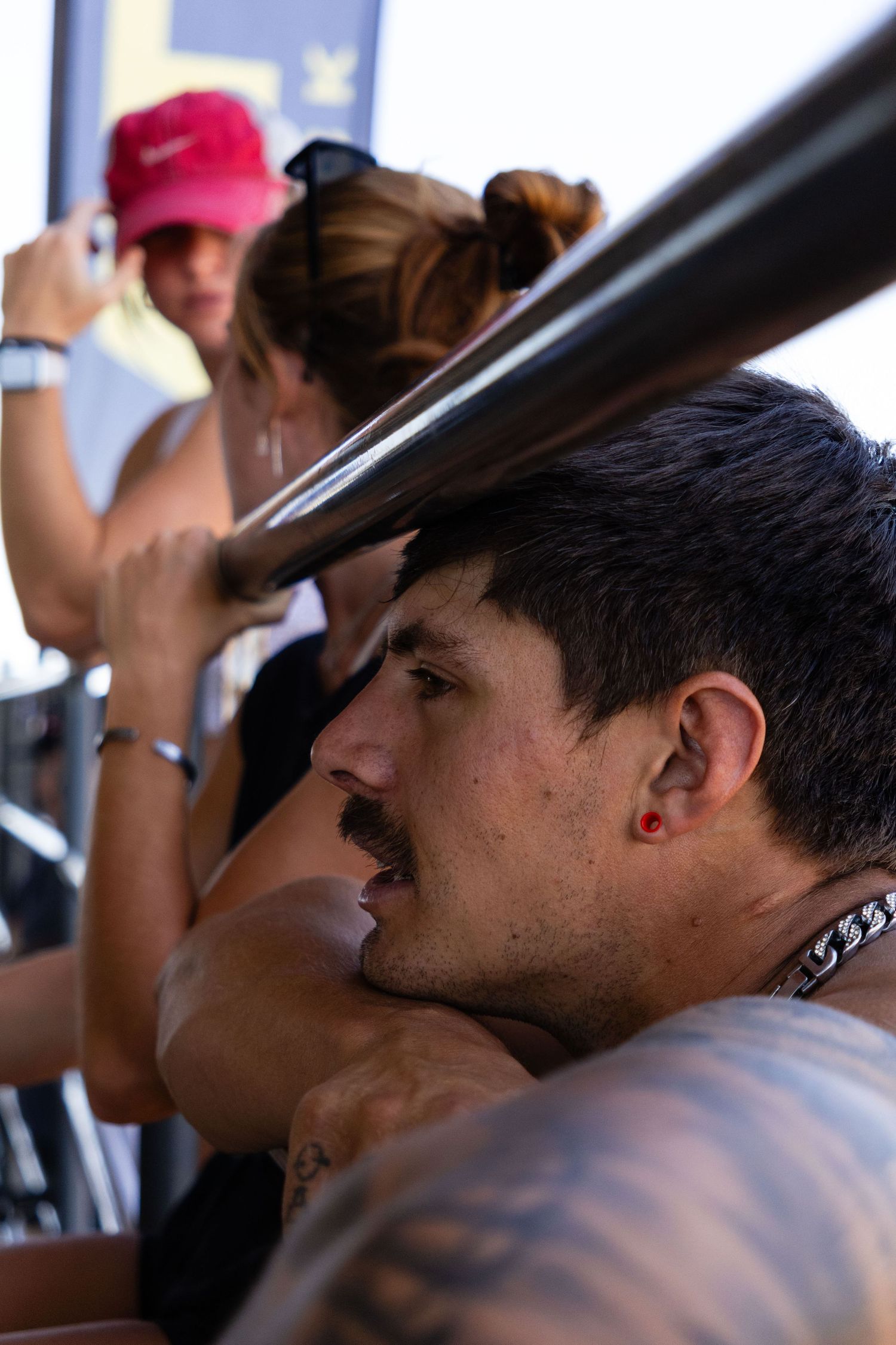 Tattooed man with red earring and mustache watching a motocross event from the spectator rail.