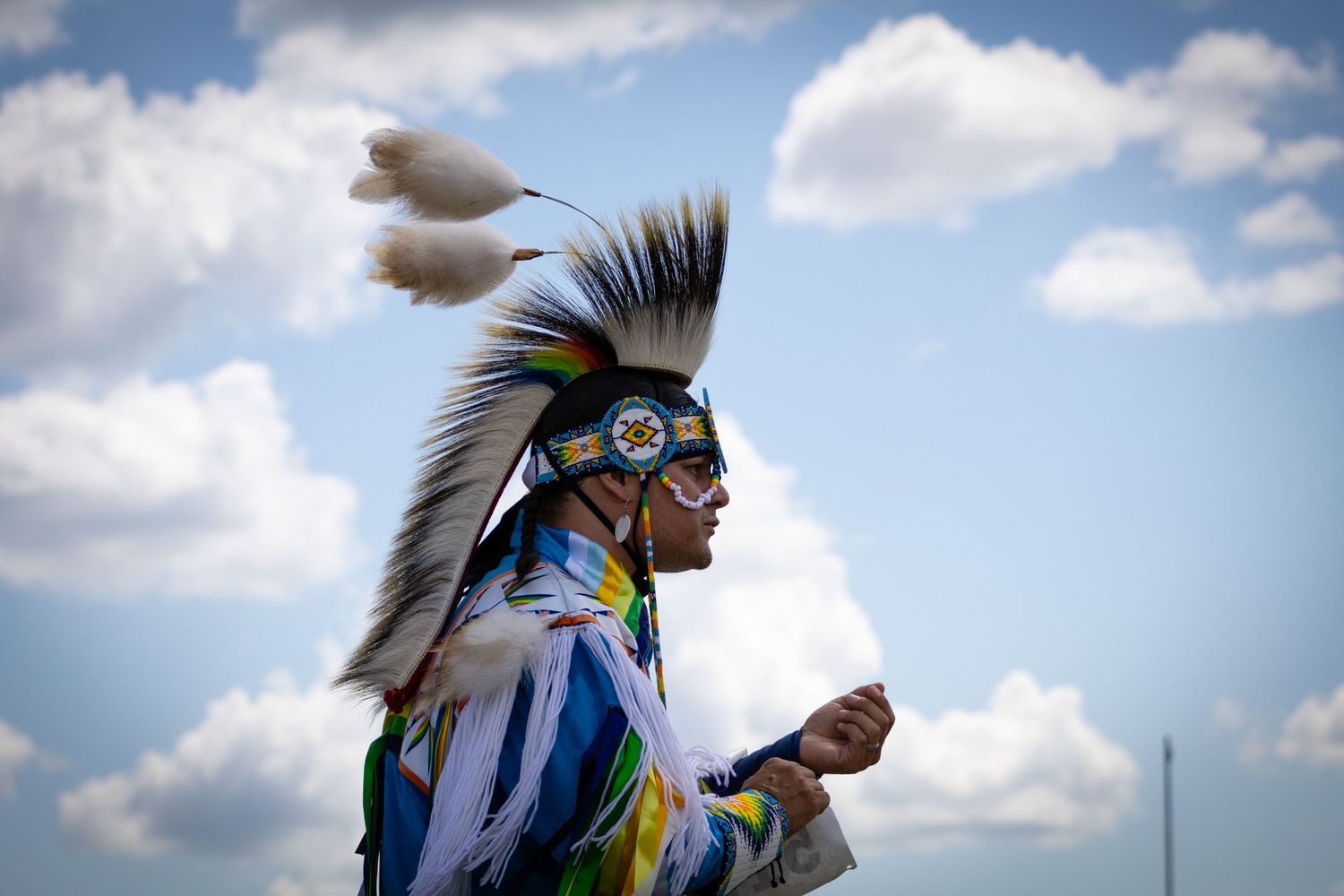 Native American man in traditional blue regalia and feathered headdress dancing against a cloudy sky.