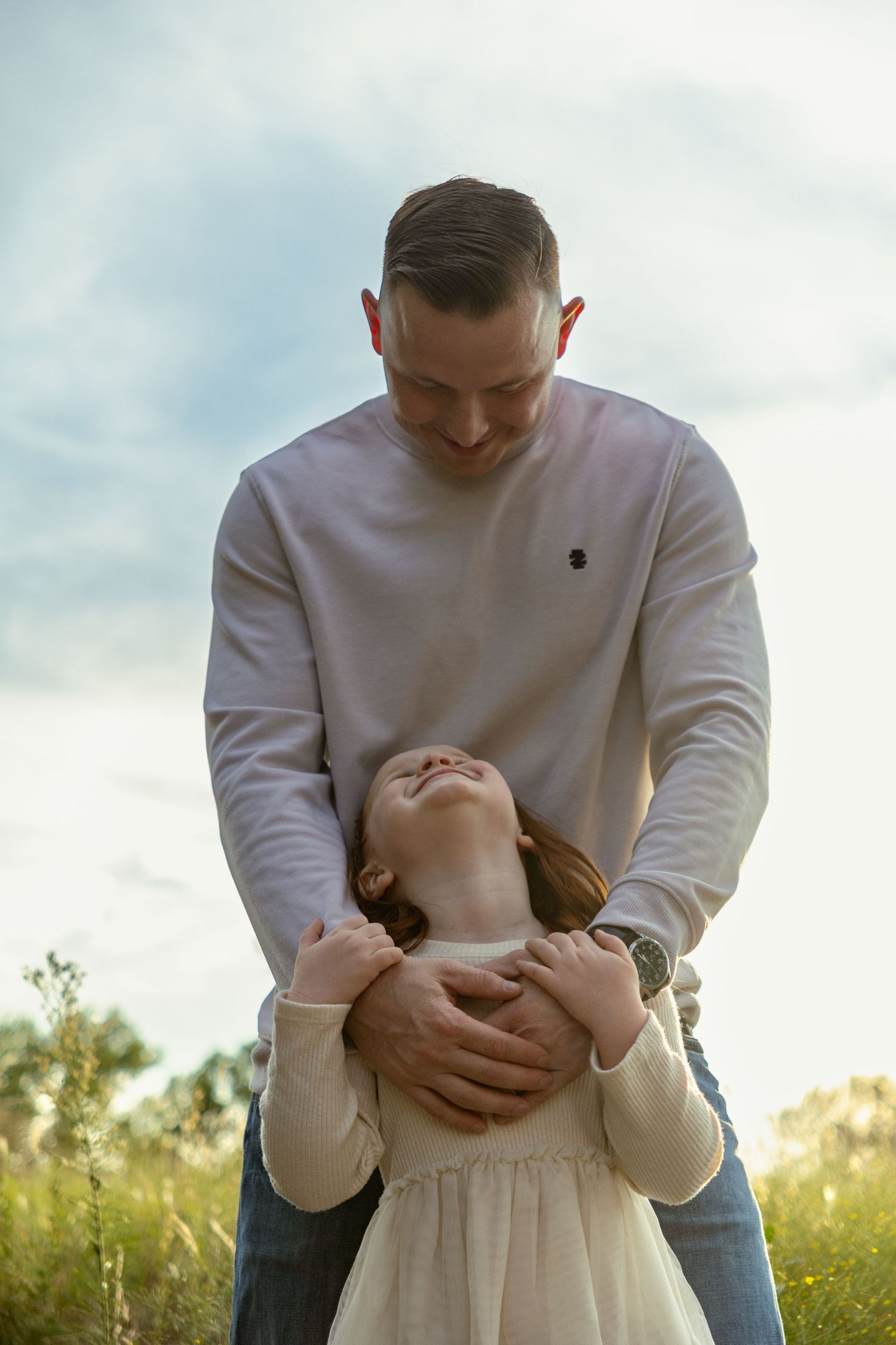 Father tenderly embracing his young daughter in a sunlit field under a cloudy sky.