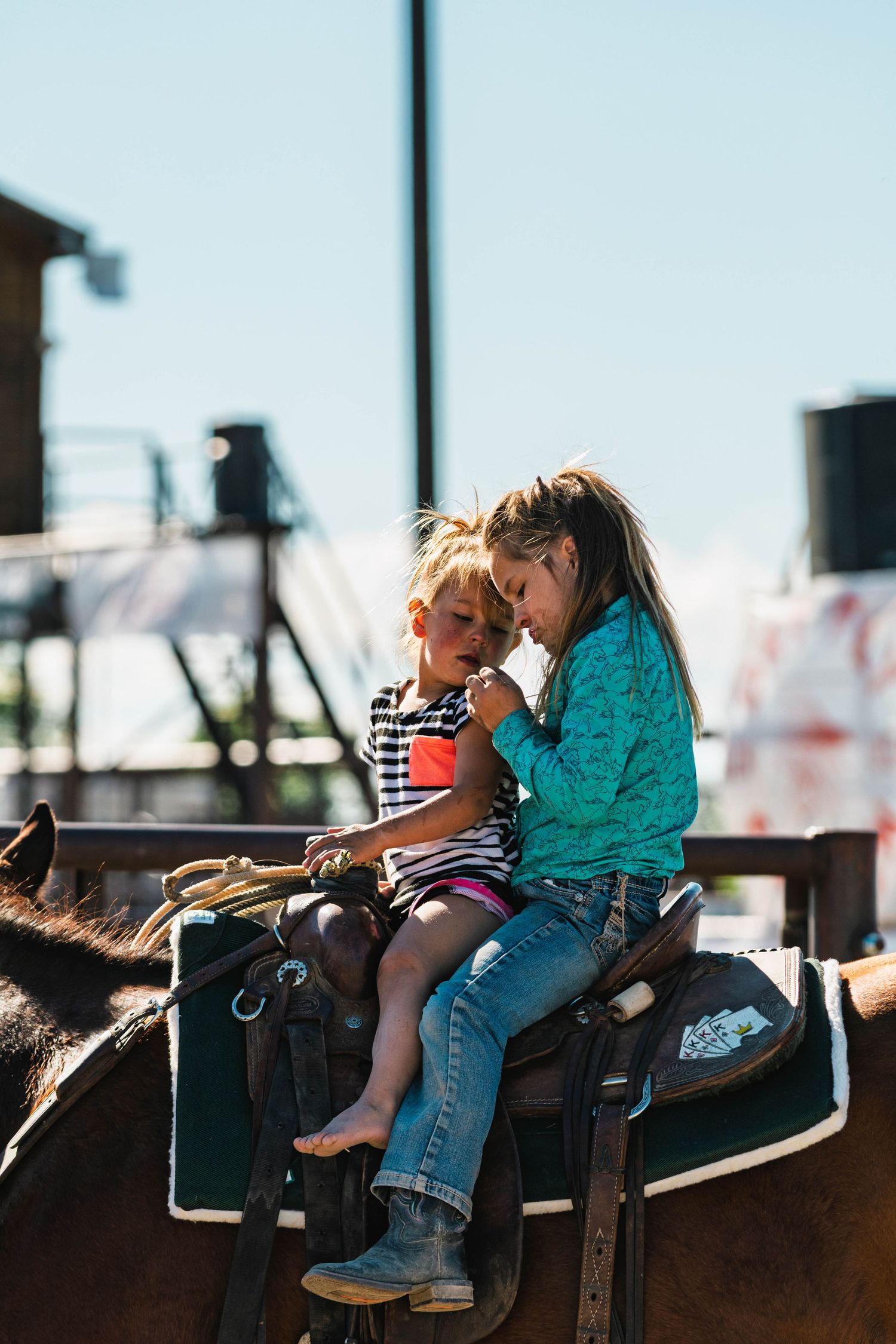 Two children sitting together on a brown horse in a saddle at an outdoor rodeo event on a sunny day.