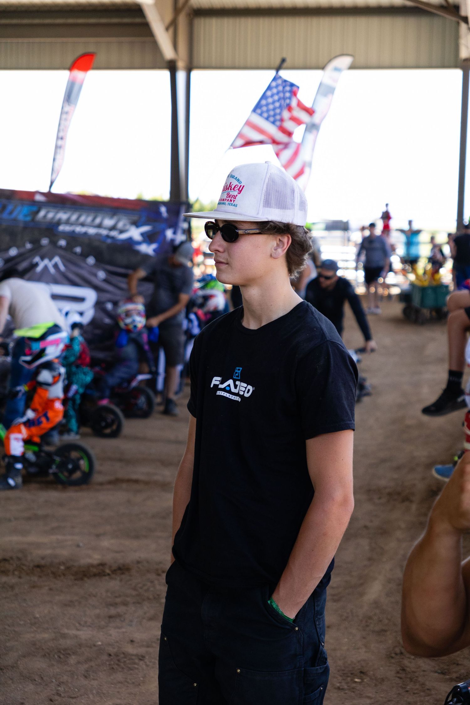 Young motocross rider in black tee and trucker hat stands at an indoor dirt bike event with American flag backdrop.