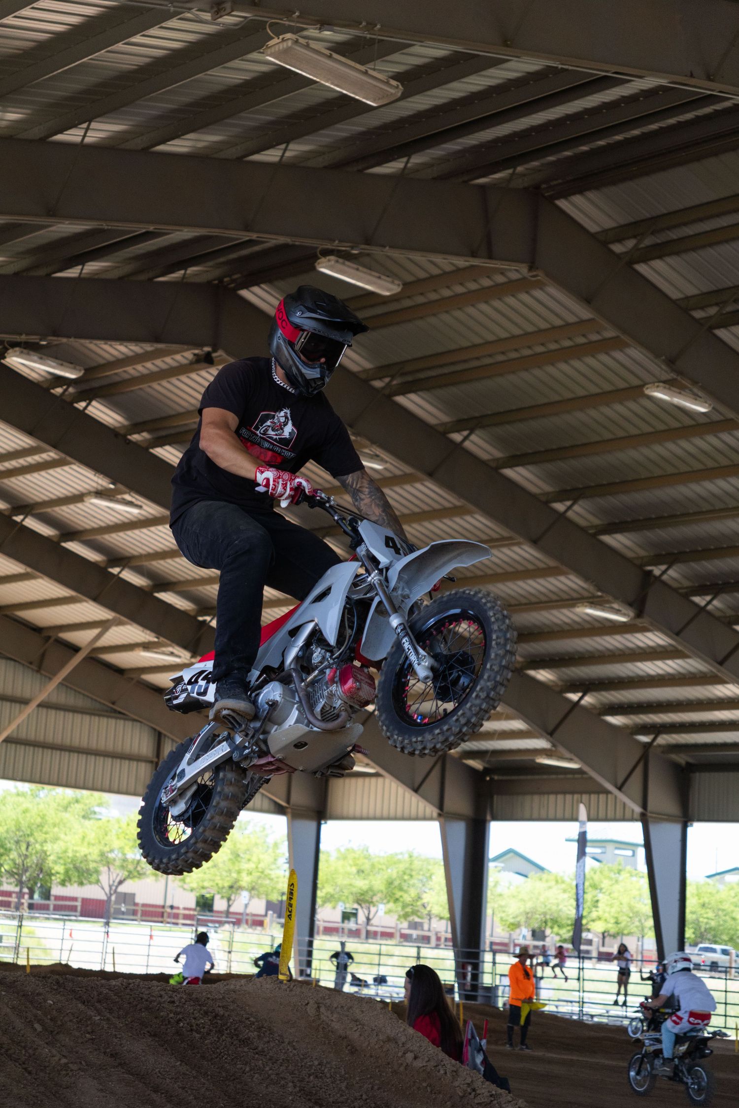 Motocross rider wearing black gear and helmet catches air on a dirt bike inside an indoor track.