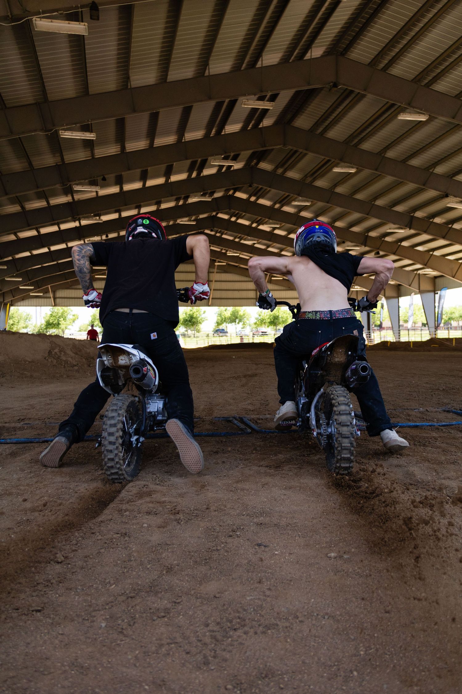 Two motocross riders perform shirtless tricks on dirt bikes inside an indoor dirt track.