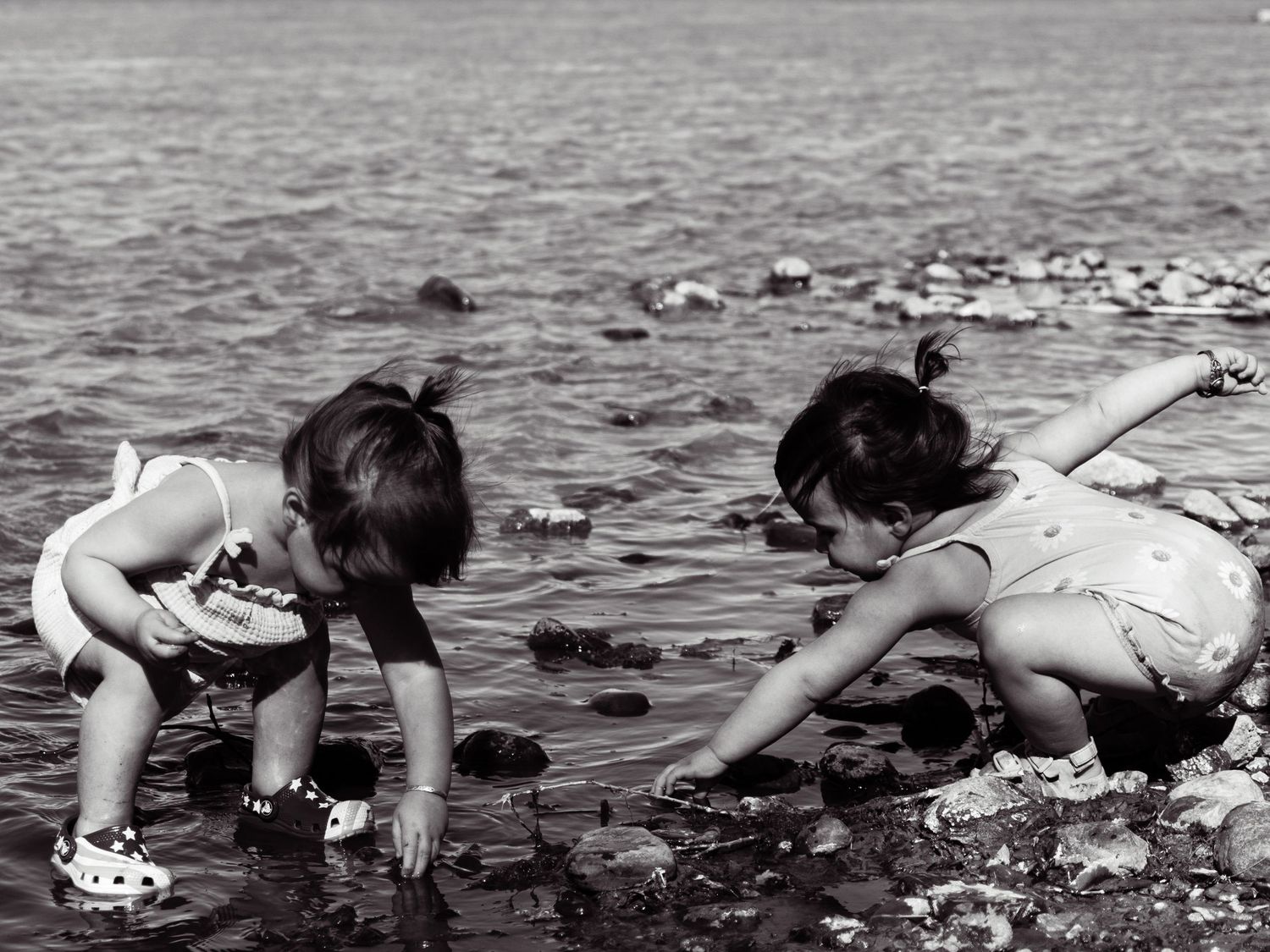Two young children crouching by a rocky shoreline exploring the water in a black and white photo.