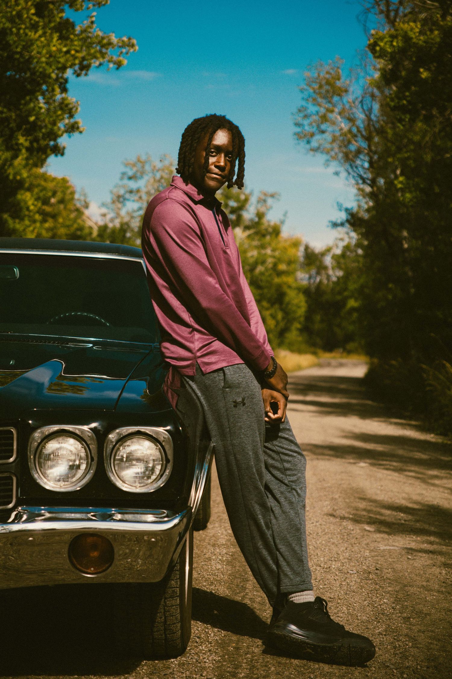 Young man in purple hoodie leaning against a vintage black muscle car on a tree-lined road.