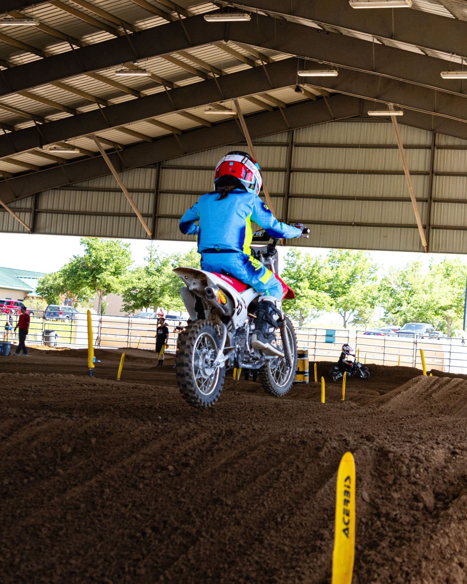 Young motocross rider in blue gear riding a dirt bike over a dirt mound in a covered arena.