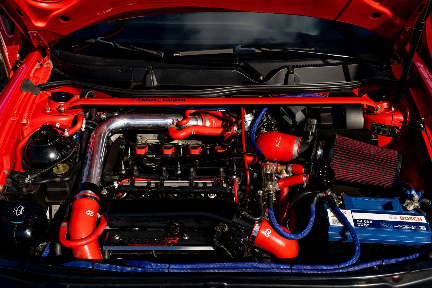 Red sports car engine bay with custom red and blue silicone hoses, cold air intake, and performance modifications.