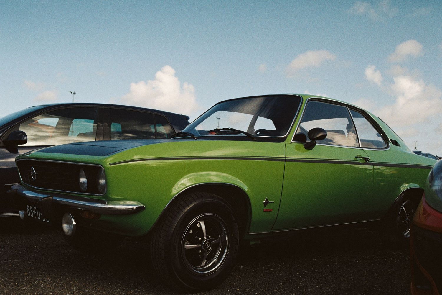 Bright green classic Opel Manta coupe parked at an outdoor vintage car show under blue sky.
