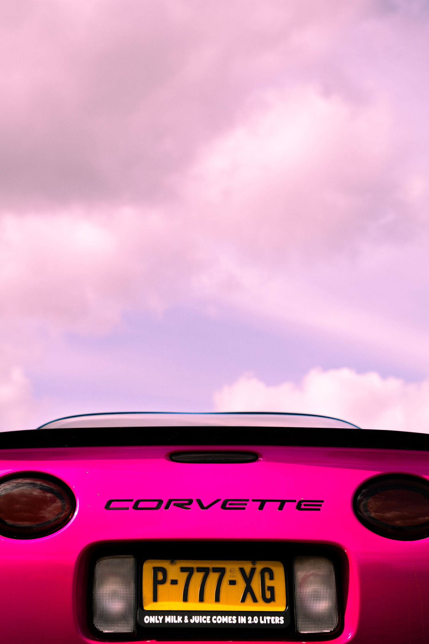 Pink Corvette sports car rear view with license plate P-777-XG against a dramatic pink and purple cloudy sky.