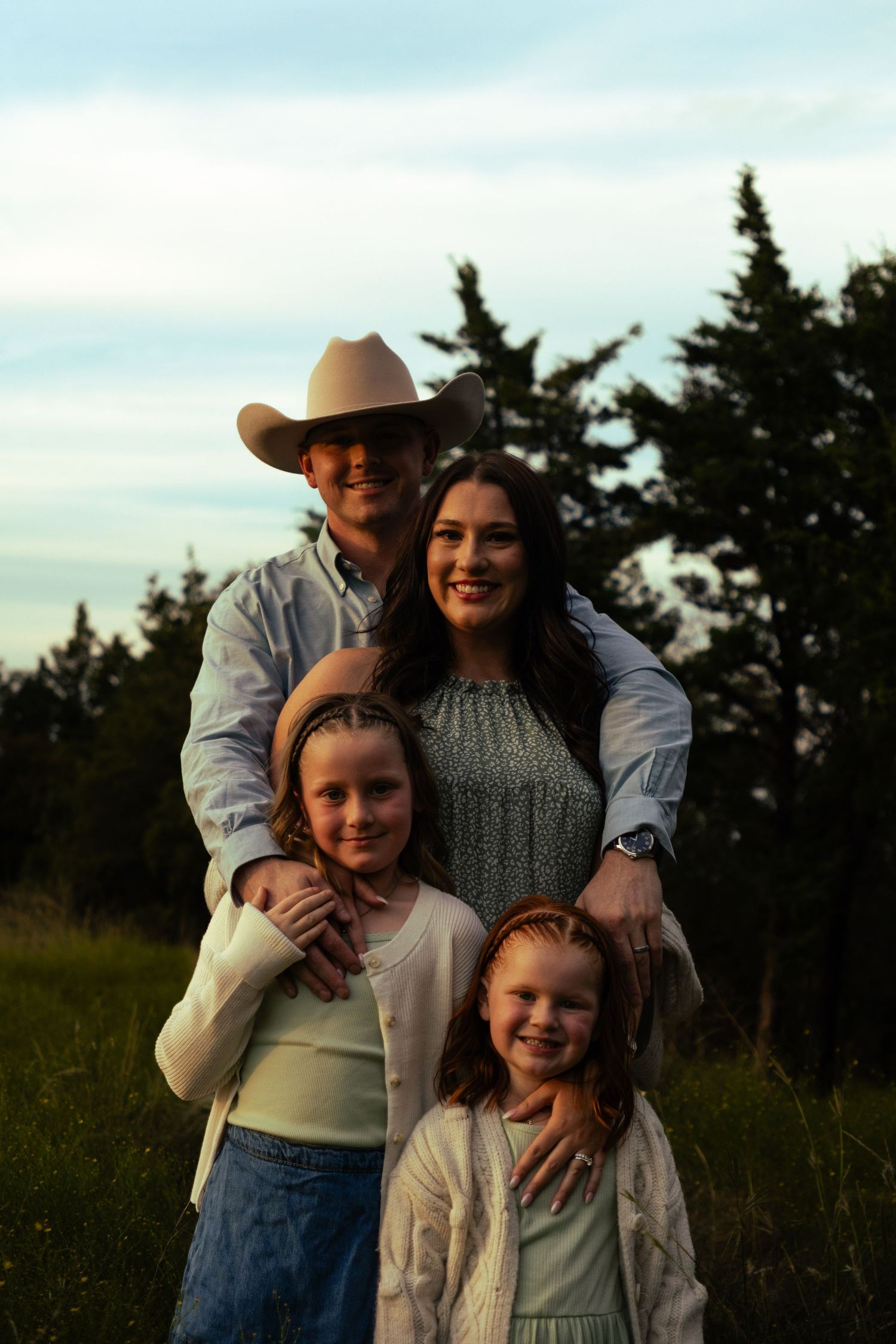 Family of four posing outdoors at golden hour with evergreen trees, father wearing a cowboy hat.