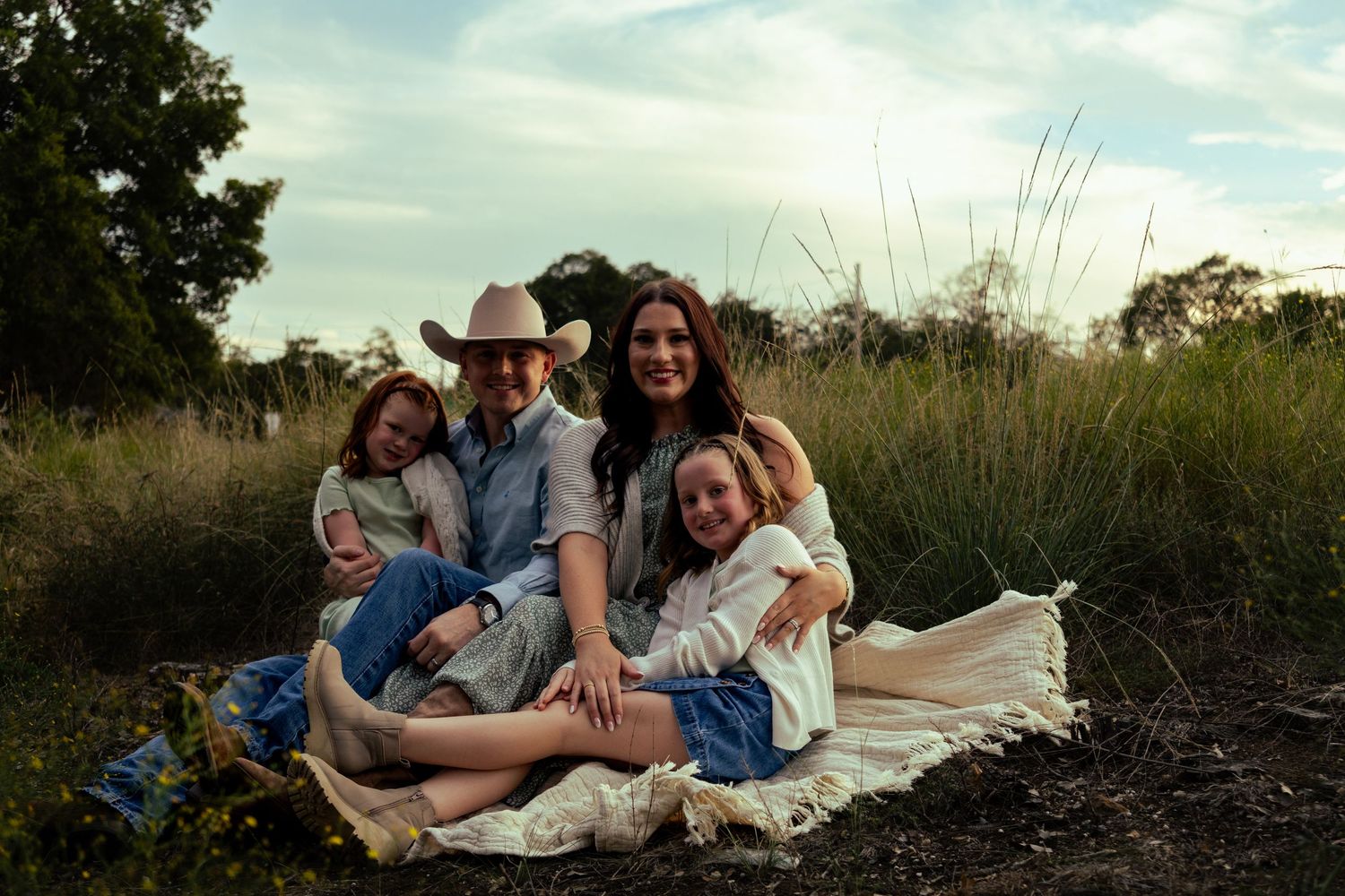 Happy family of four sitting on a blanket in a grassy meadow at golden hour, father in cowboy hat.