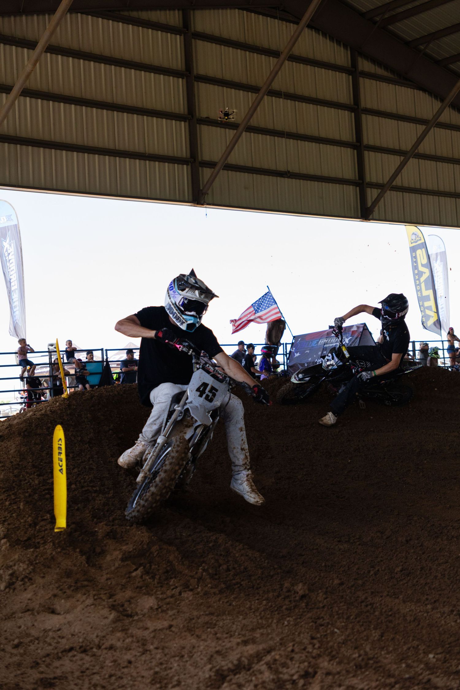 Two motocross riders navigate dirt berms inside covered arena with American flag visible in background.