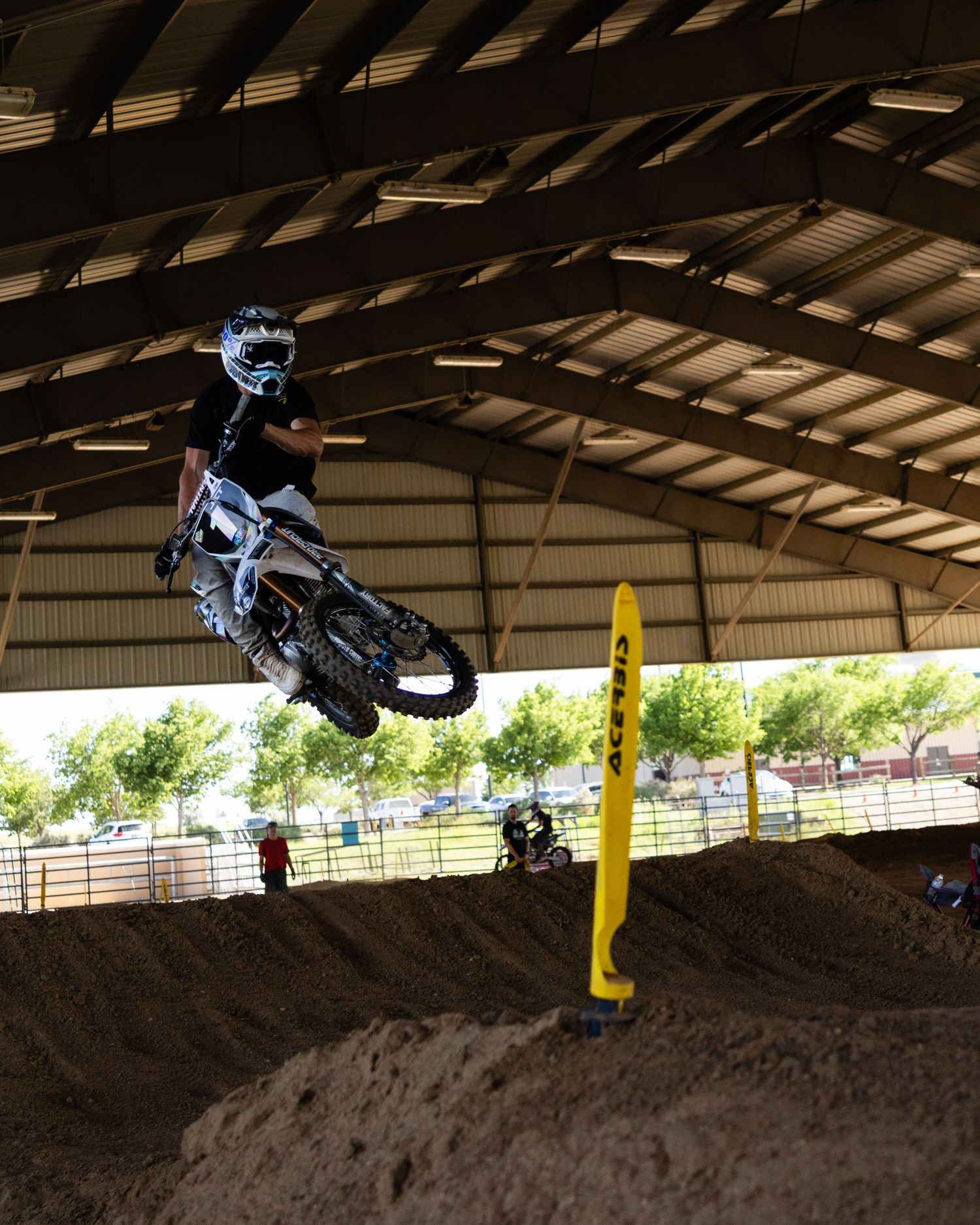Motocross rider catching air on a dirt jump inside a covered indoor track facility.