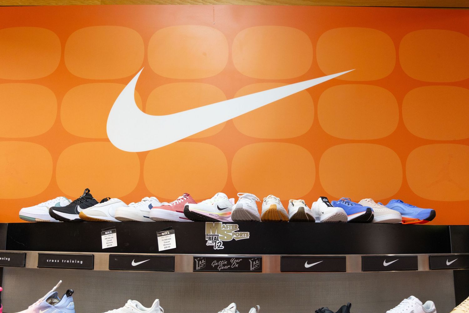 Nike sneakers displayed on a shelf below the iconic white Nike swoosh logo on an orange wall in a retail store.