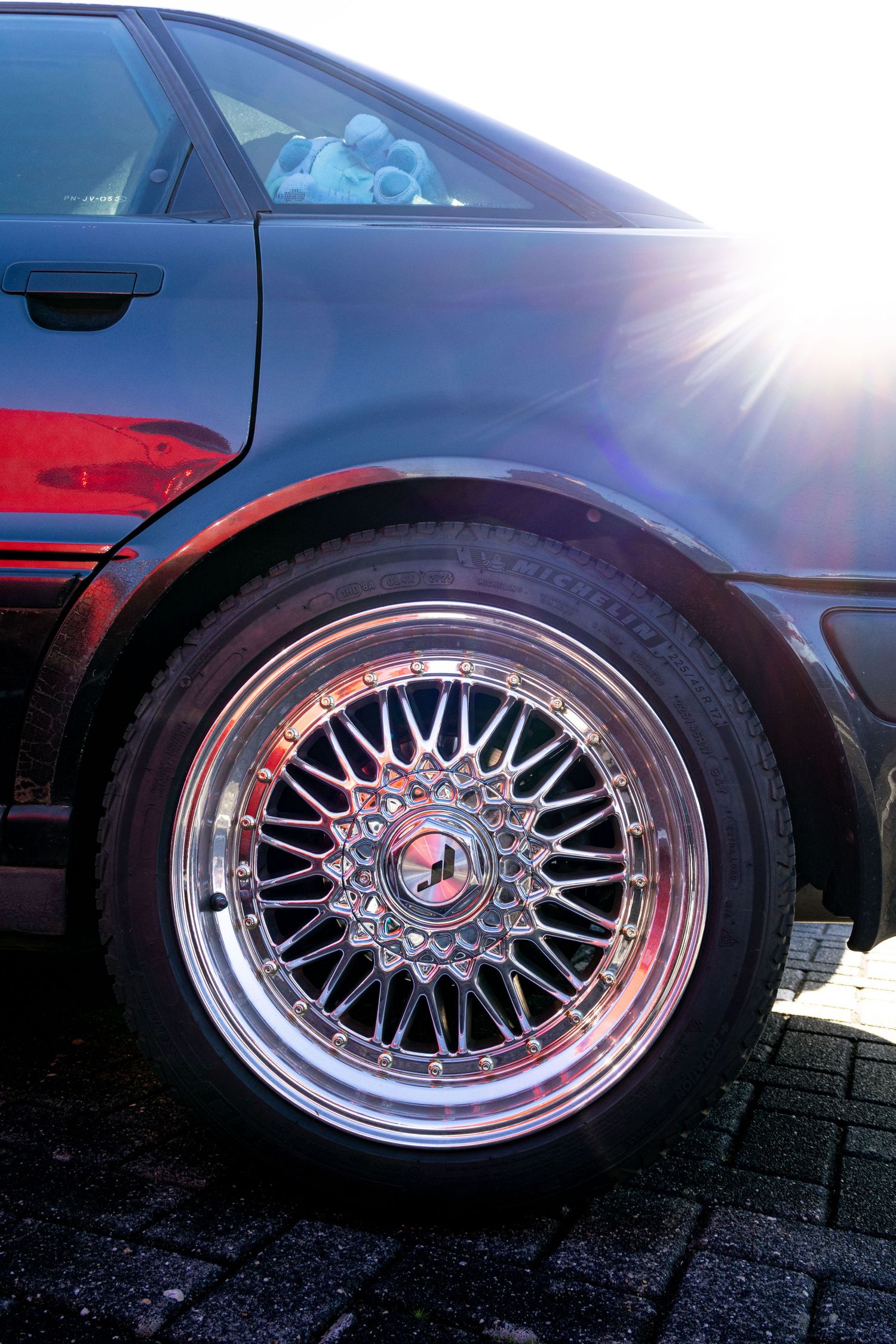 Close-up of a polished chrome BBS mesh wheel on a black luxury car with sunburst flare in background.