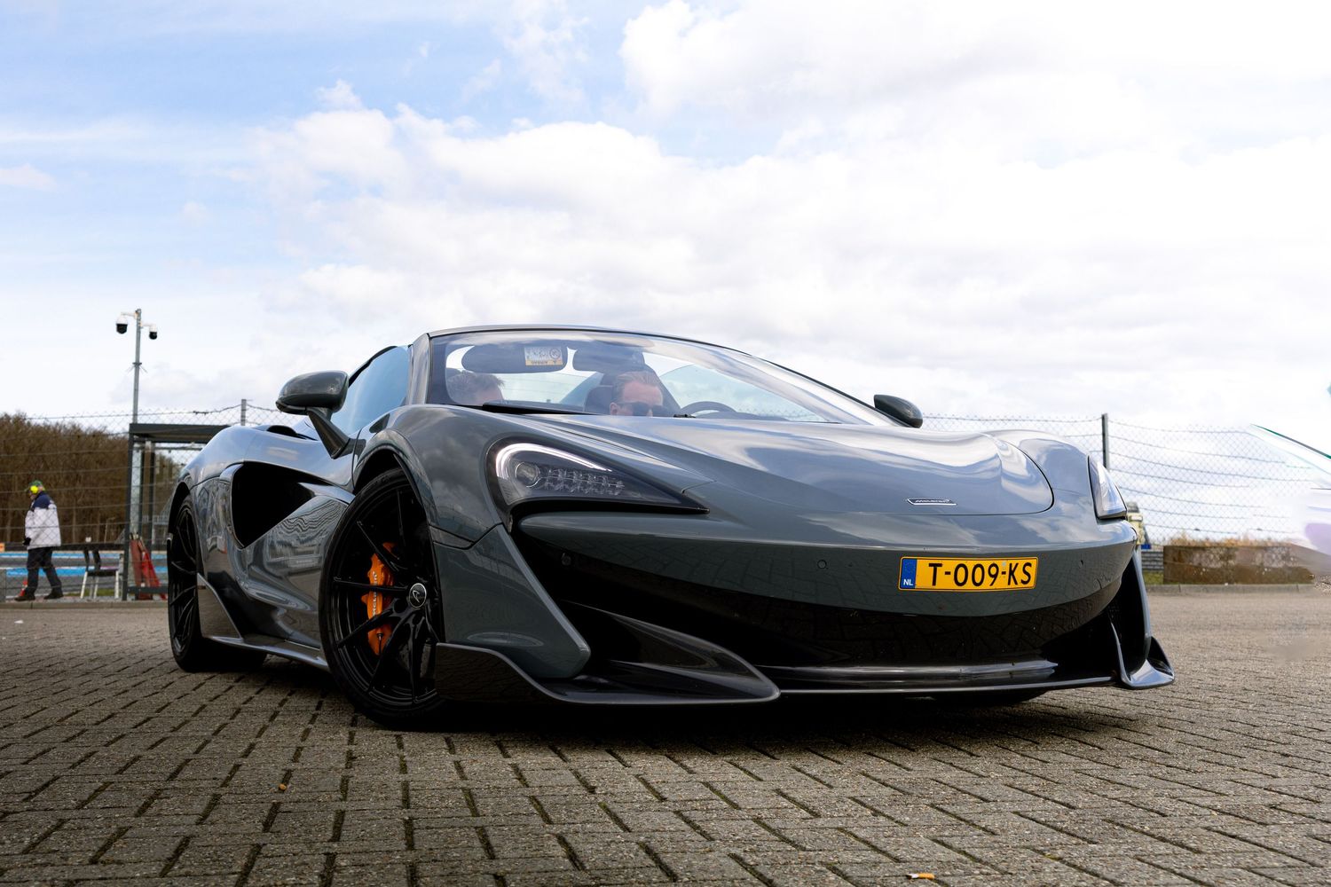 Gray McLaren 600LT supercar with Dutch license plate parked on a lot under cloudy skies.