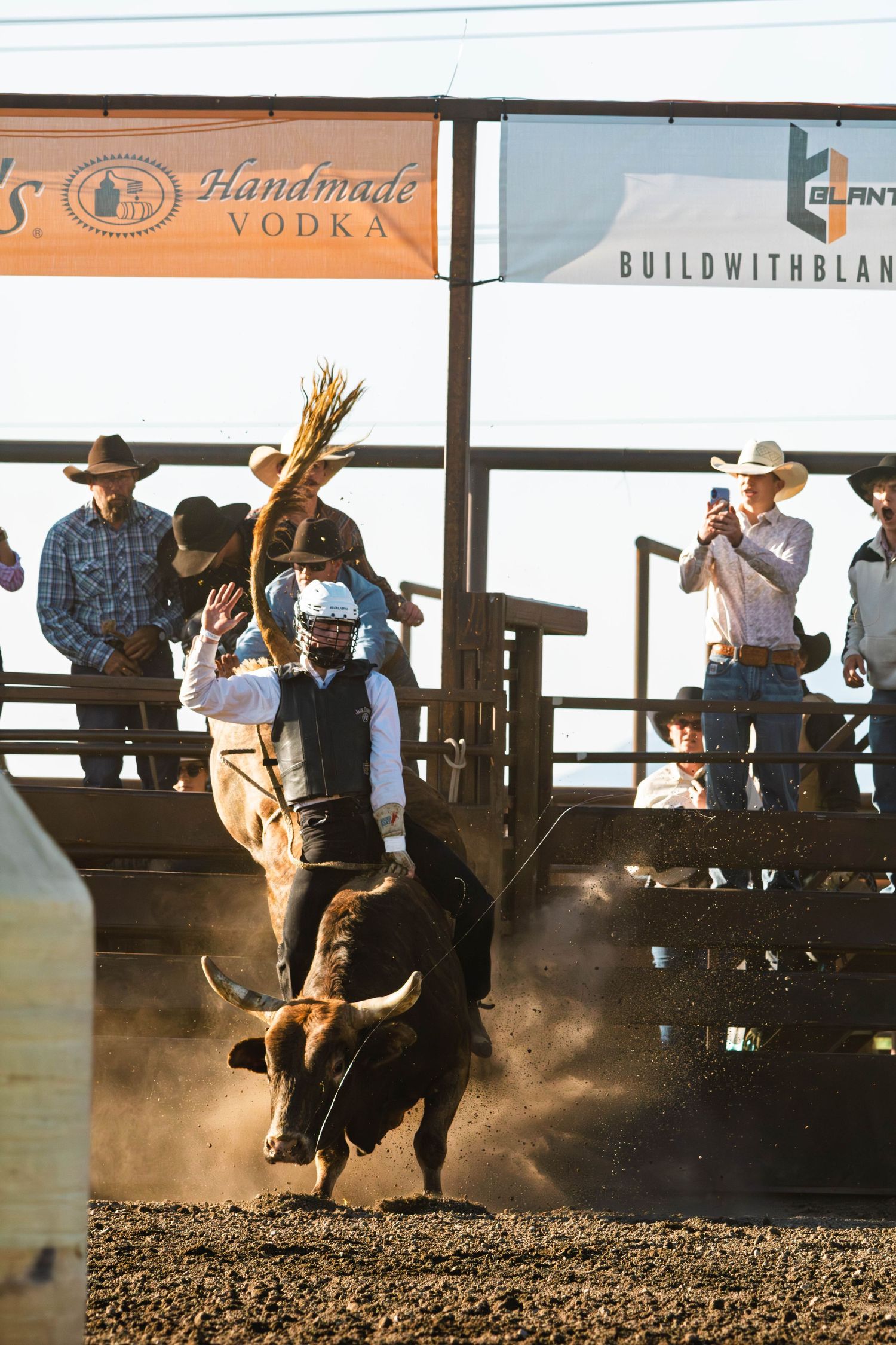 A bull rider competes at a rodeo arena while spectators watch from behind the fence, with sponsor banners visible.