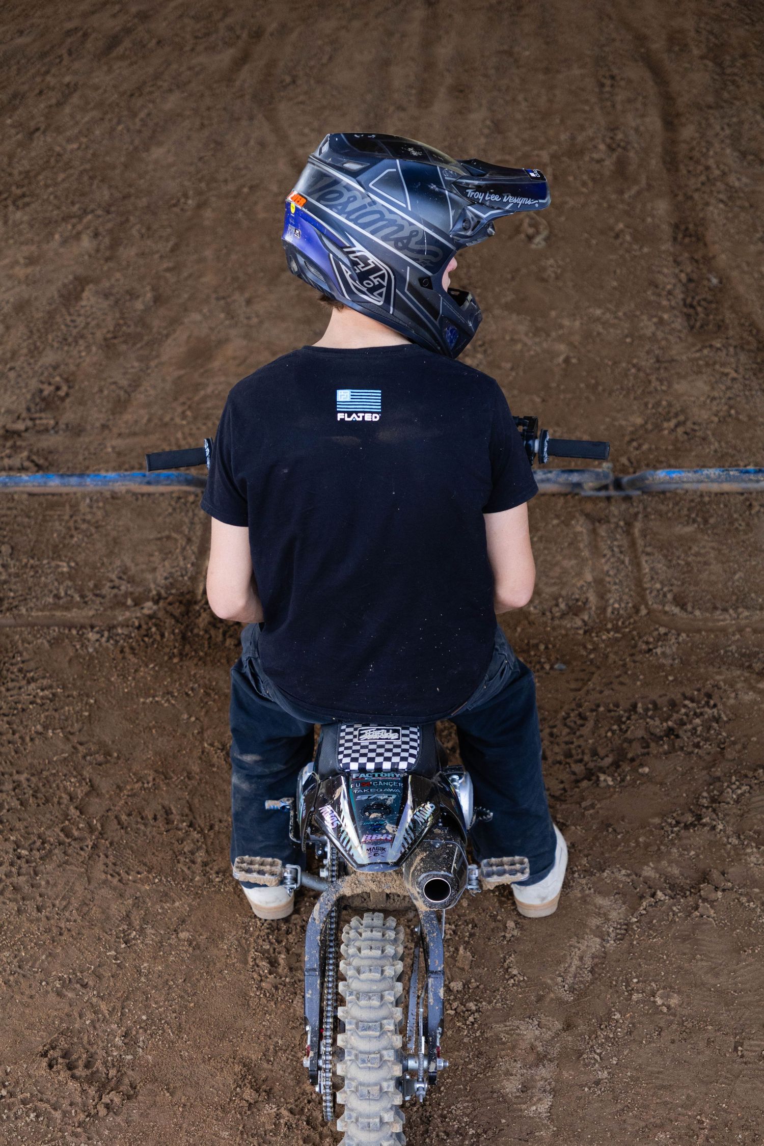 Rear view of a young motocross rider sitting on a dirt bike on an indoor dirt track.