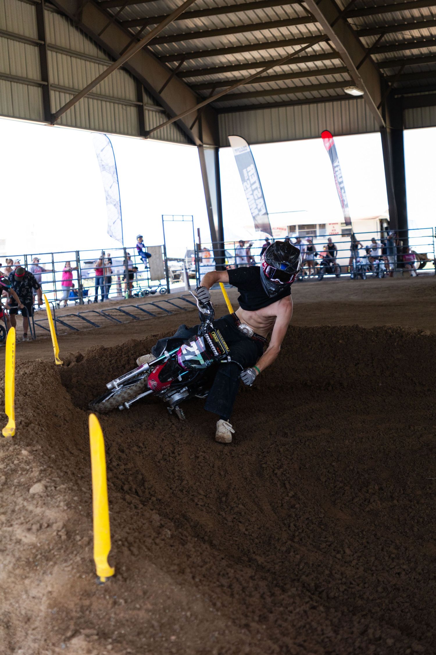 Motocross rider crashes on a dirt berm inside an indoor arena with spectators watching.