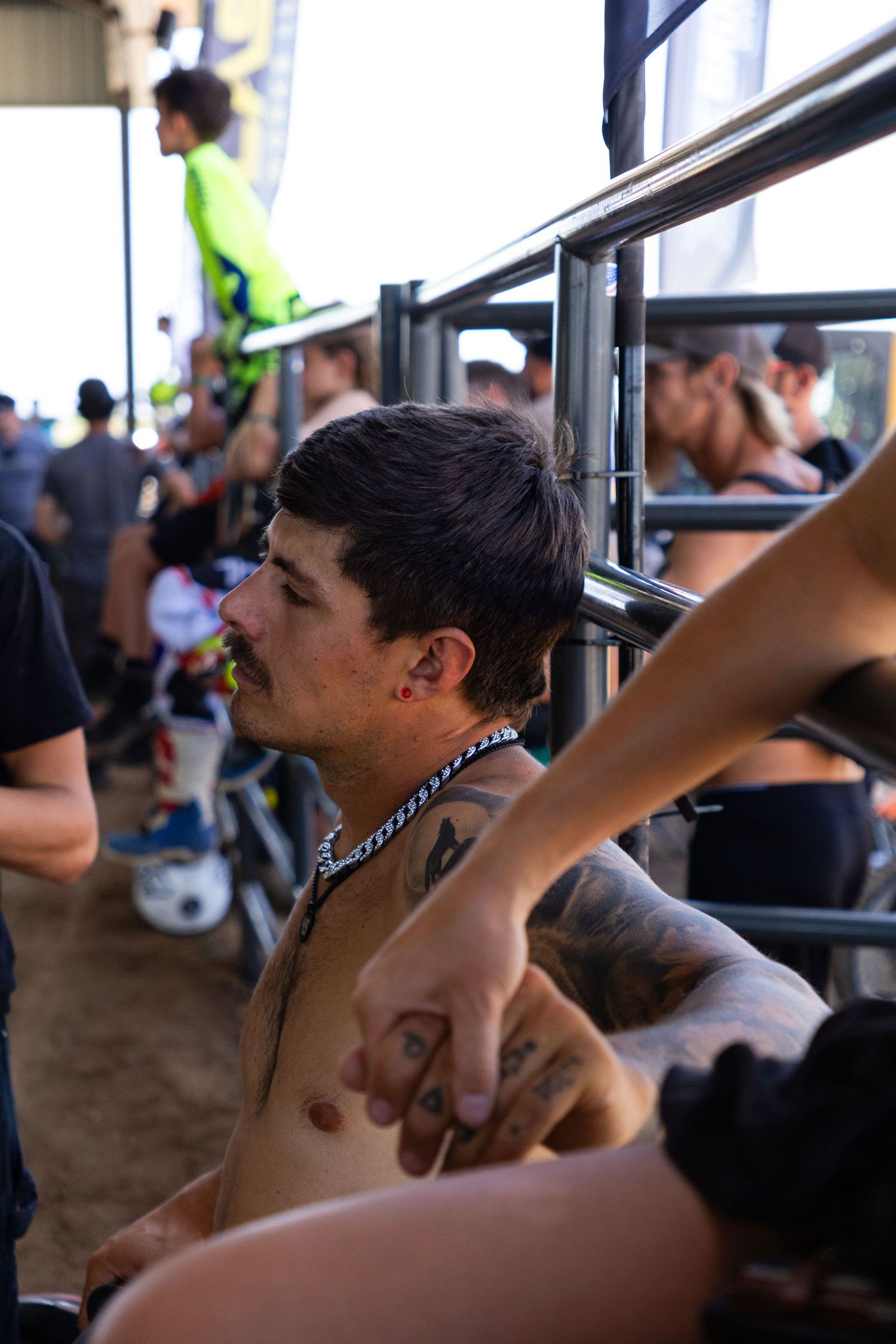 Tattooed shirtless man with chain necklace sits in bleachers at an outdoor motocross racing event.