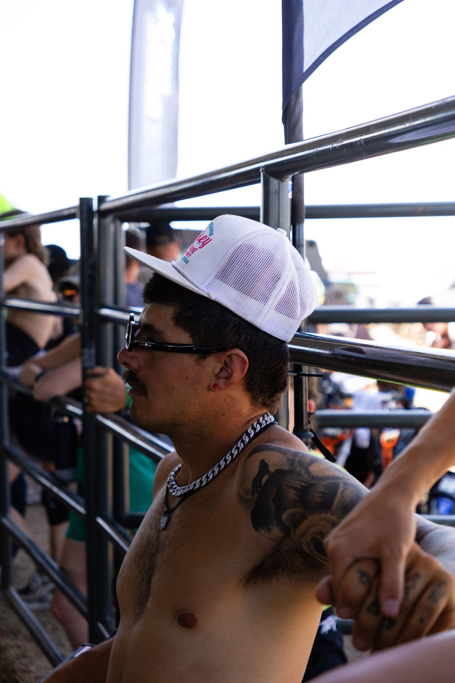 Tattooed shirtless man wearing white trucker cap and chain necklace watches motocross event from bleachers.