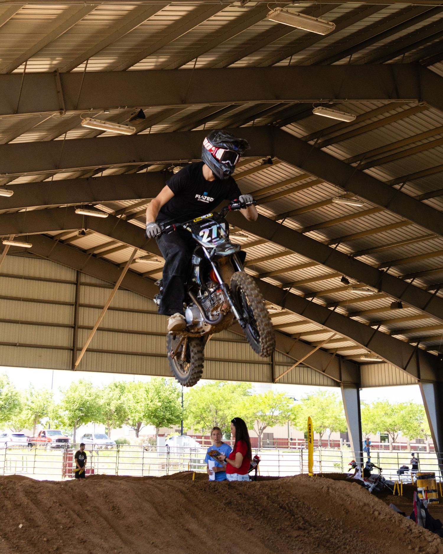 Motocross rider launching off a dirt jump inside a covered arena during an indoor supercross event.