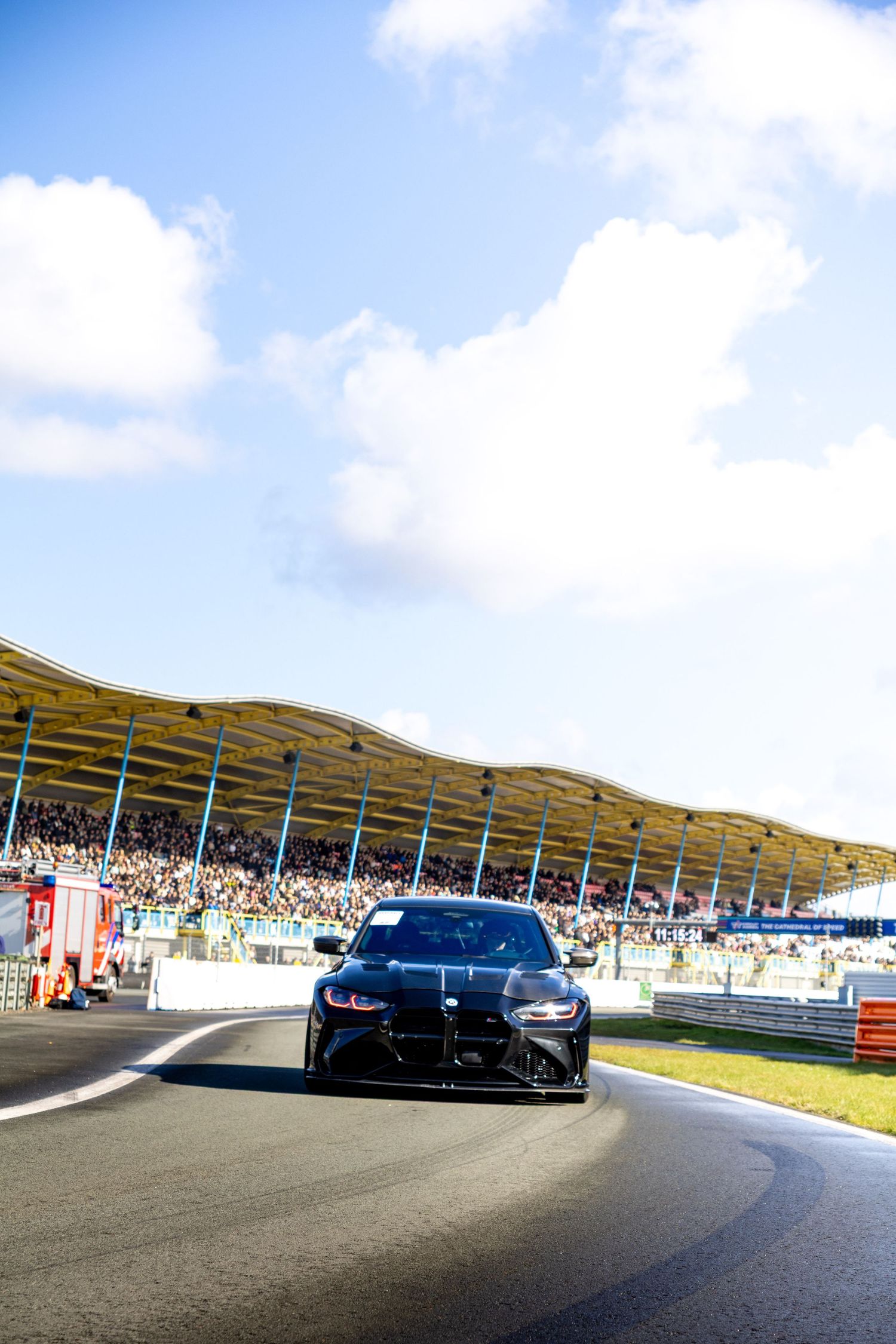 Black BMW M4 racing on a circuit track with packed grandstands and blue sky at a motorsport event.