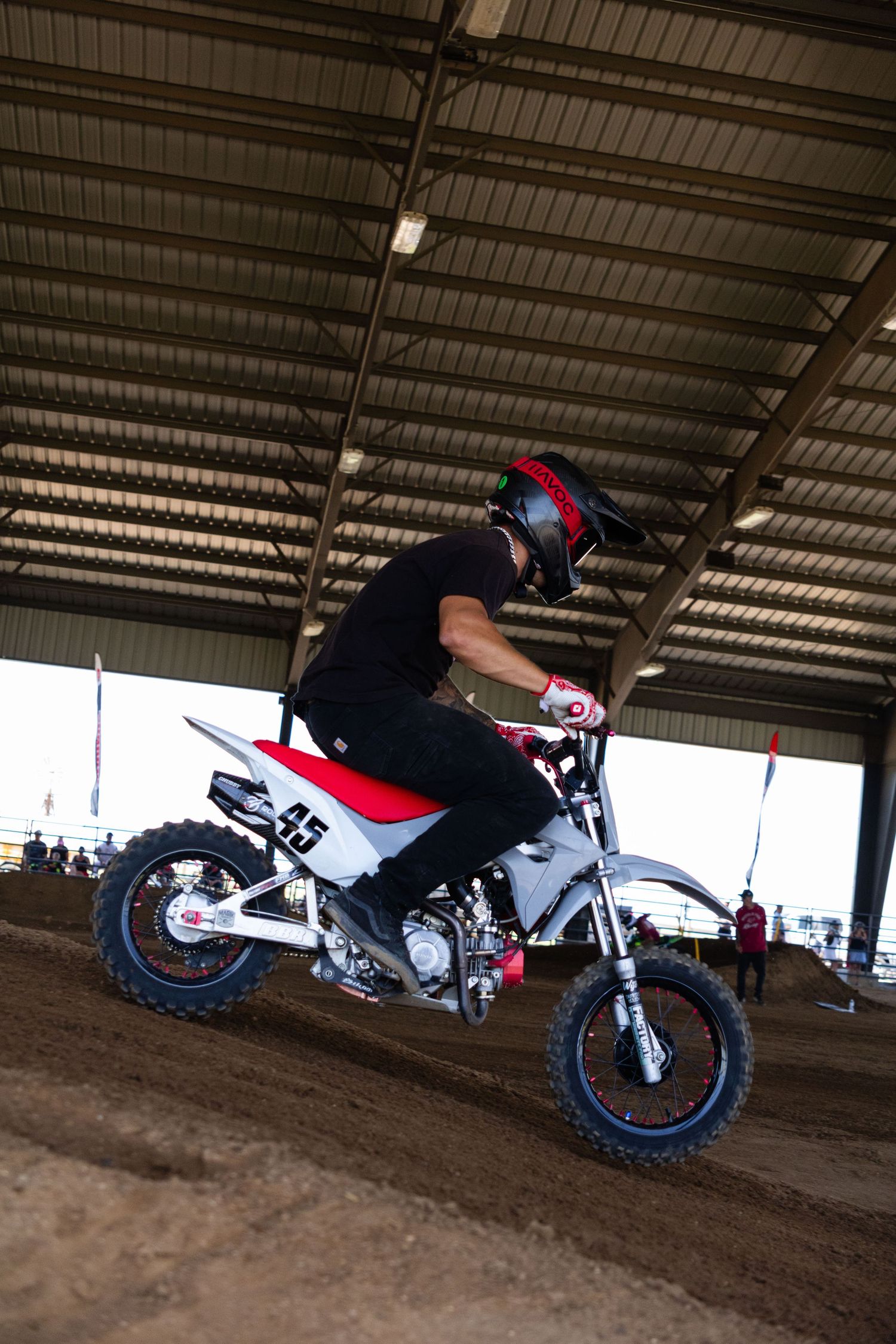 Motocross rider on red and white dirt bike accelerating on indoor dirt track in arena.