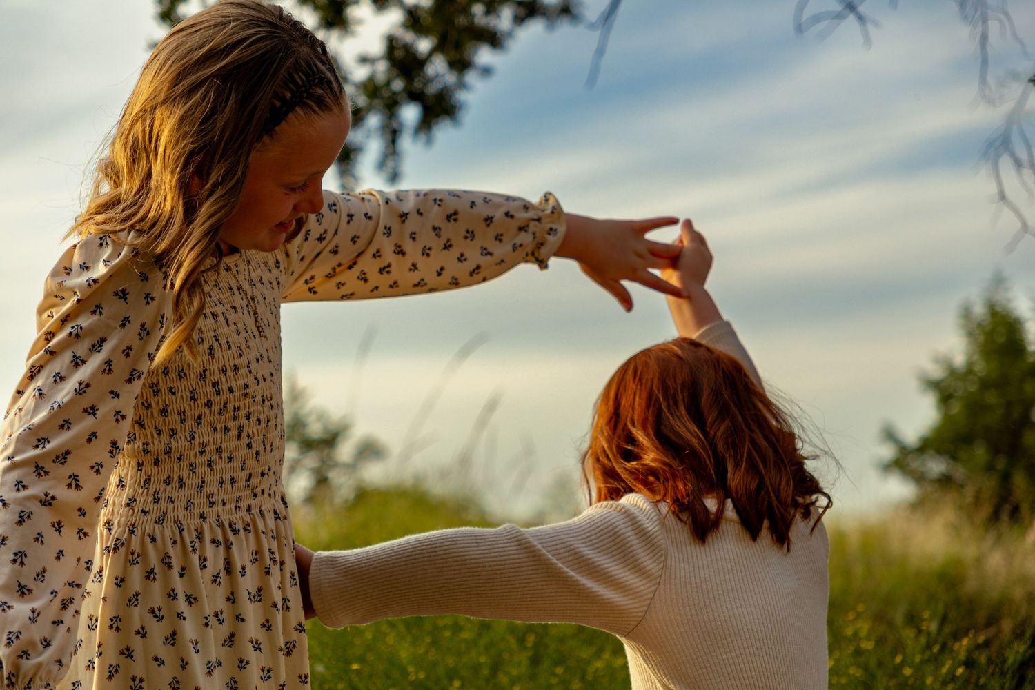 Two young girls dancing together outdoors in a field with a cloudy sky backdrop.