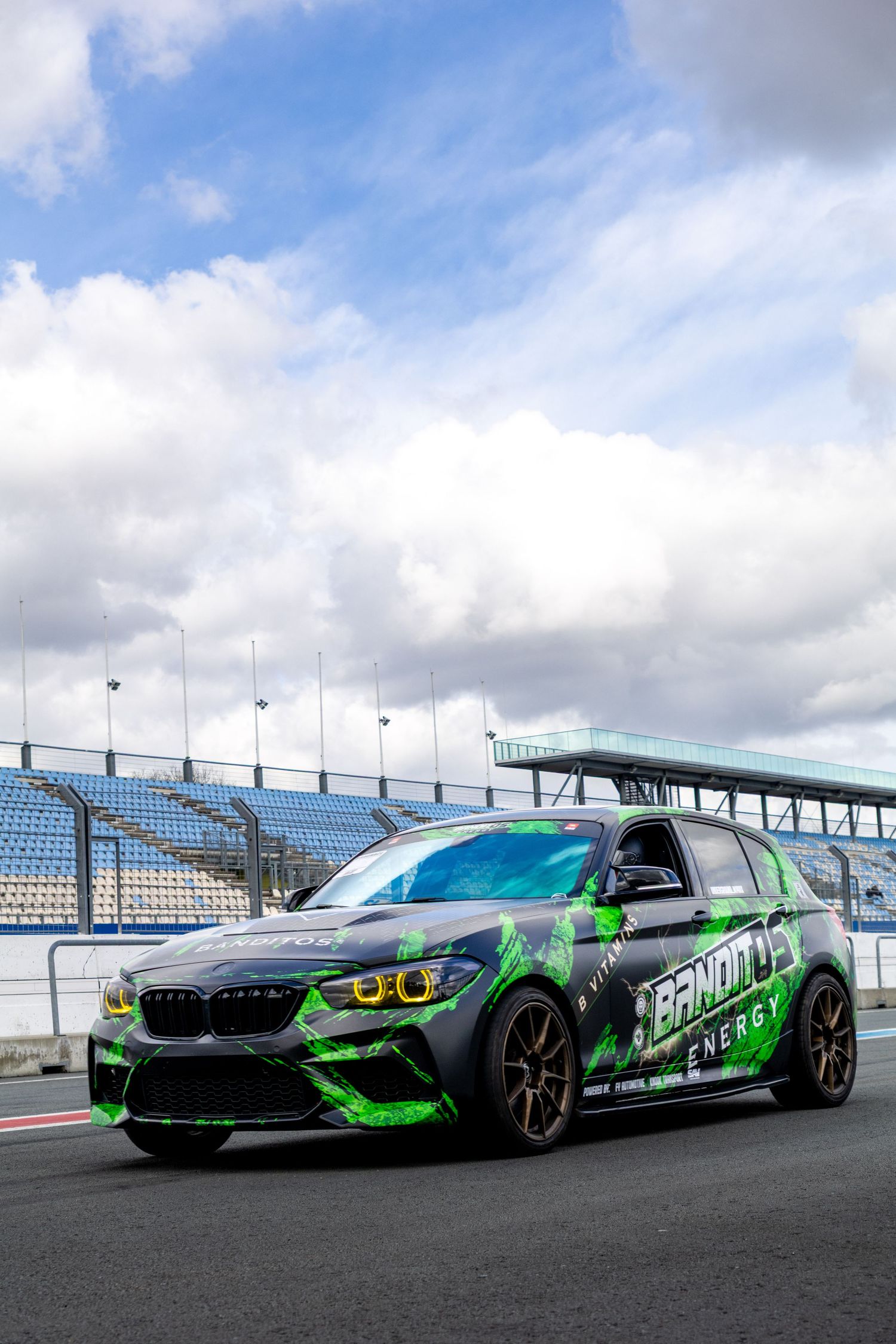 BMW M2 race car with green Bandito Energy livery parked on a racing circuit with grandstands in the background.