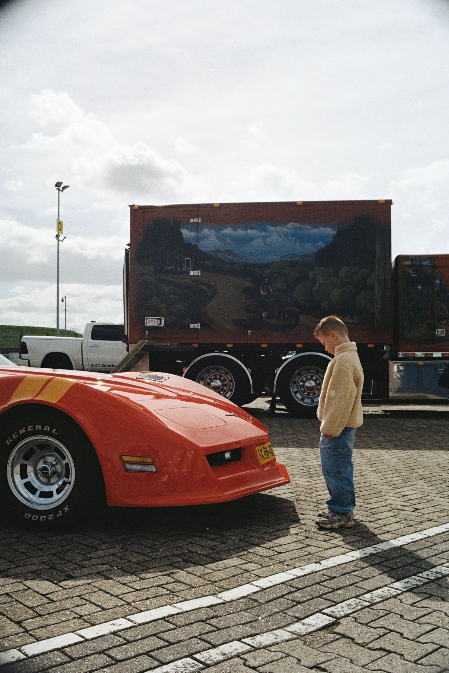 Child standing beside a red Corvette sports car in a parking lot with a scenic mural truck in the background.