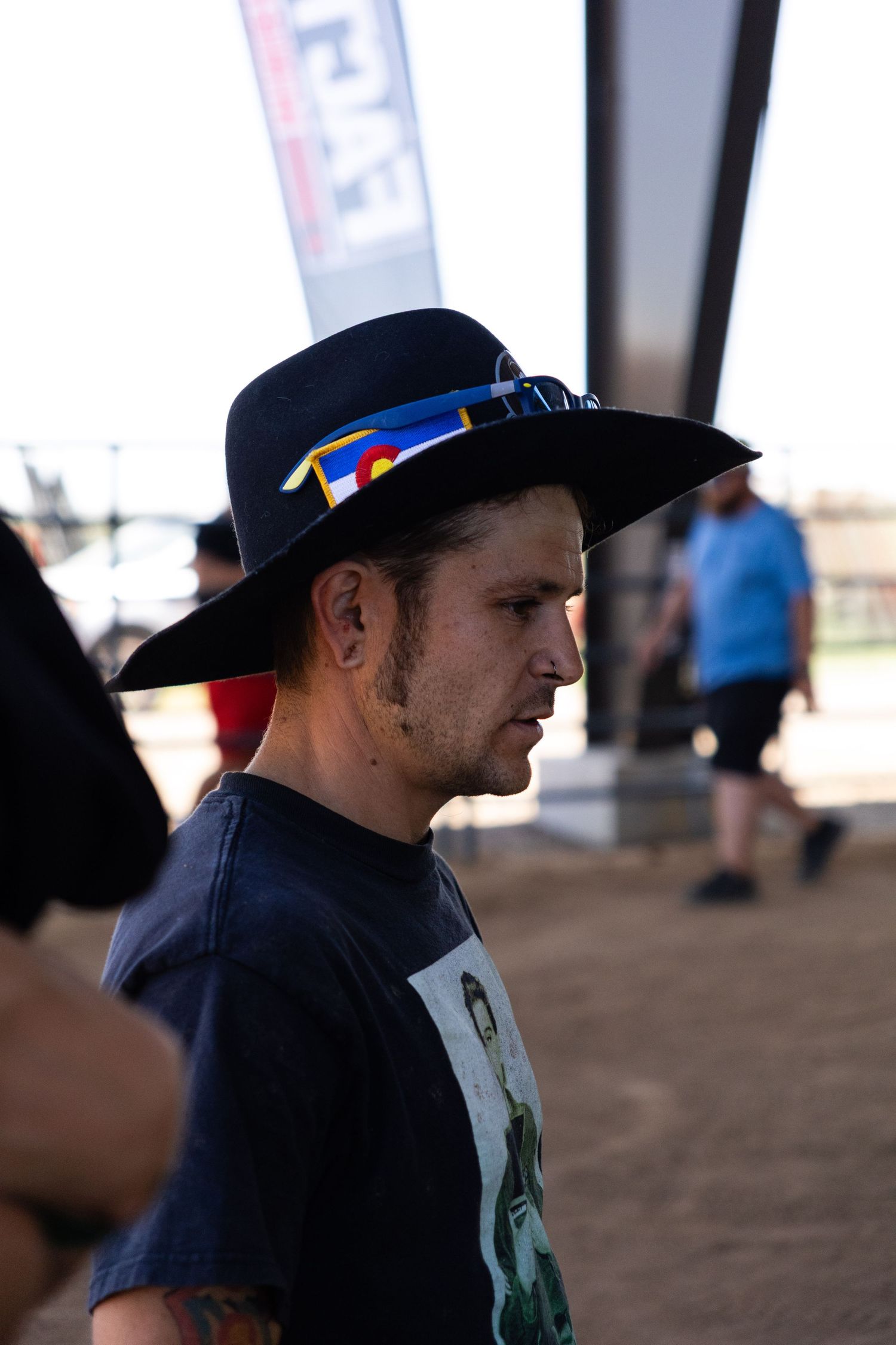 Man in black wide-brim hat with Colorado flag band standing at an outdoor motocross event.