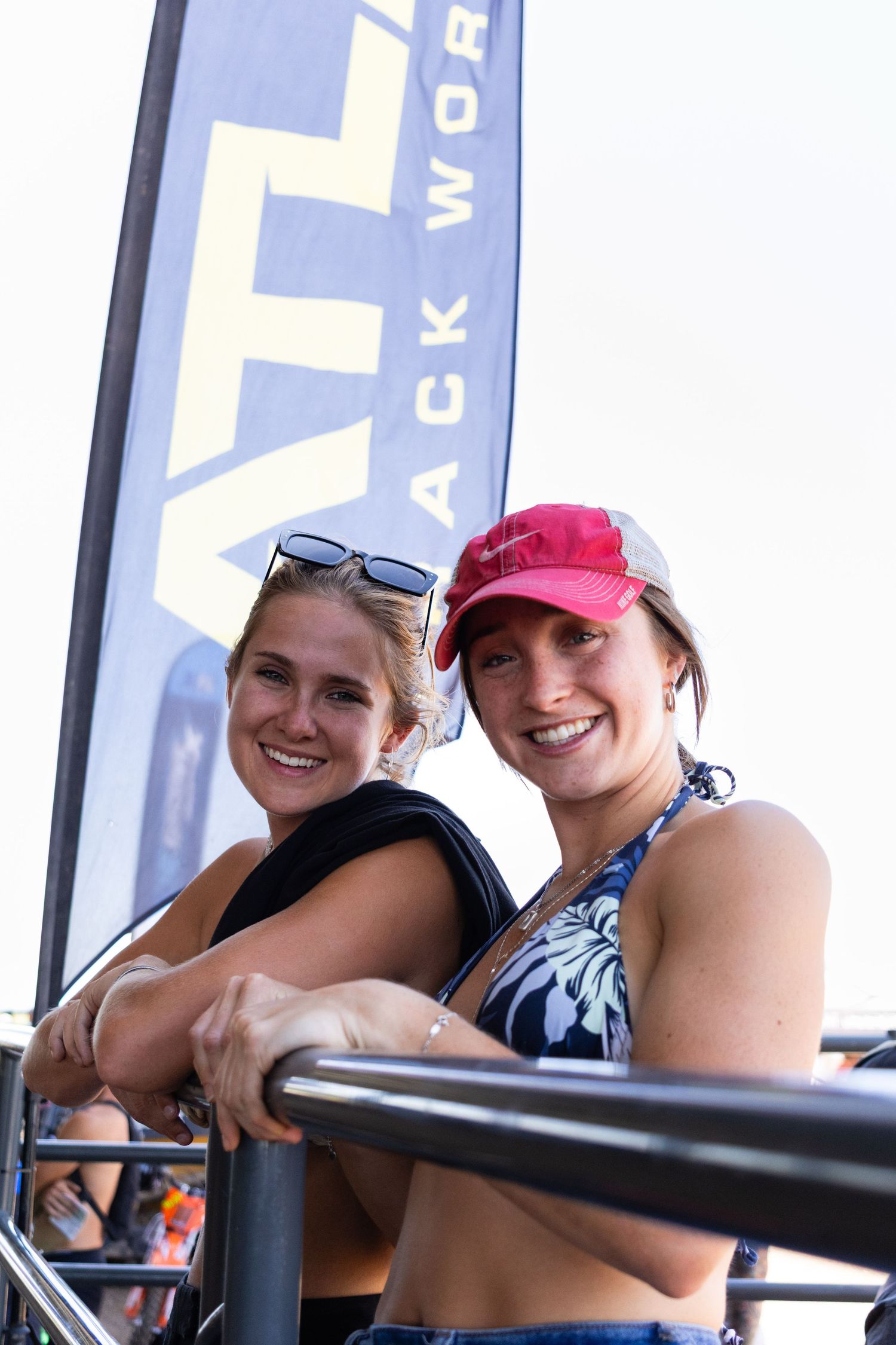 Two smiling women leaning on a railing at an outdoor motocross event with an Atlas banner behind them.