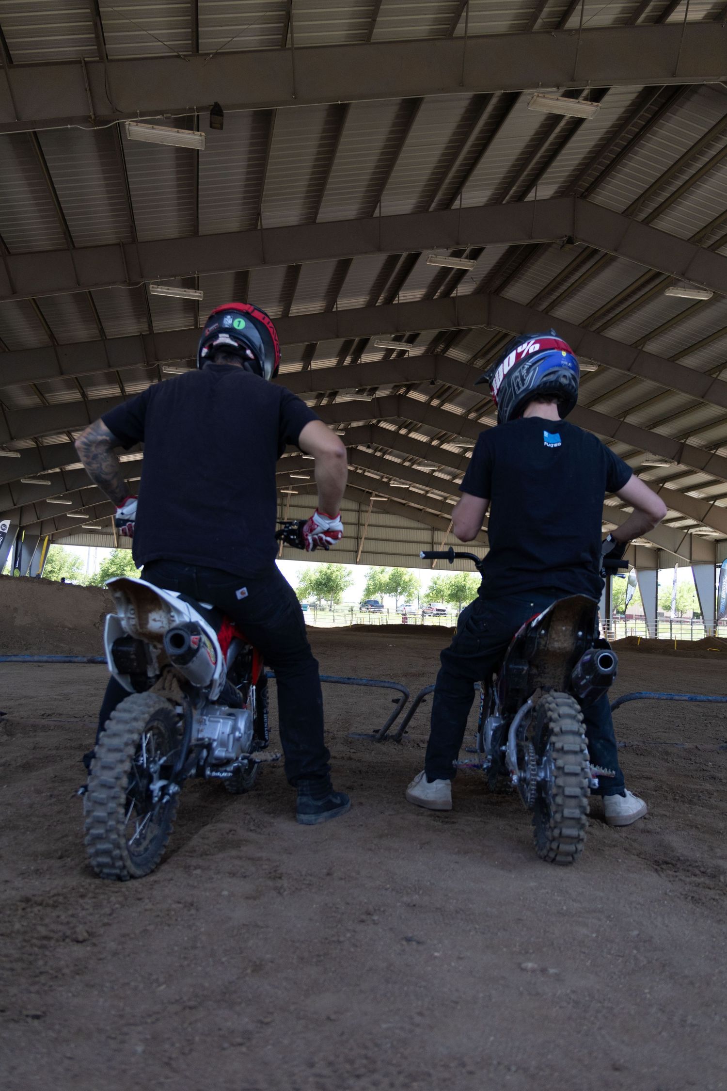 Two helmeted riders stand beside their dirt bikes inside a covered motocross arena.