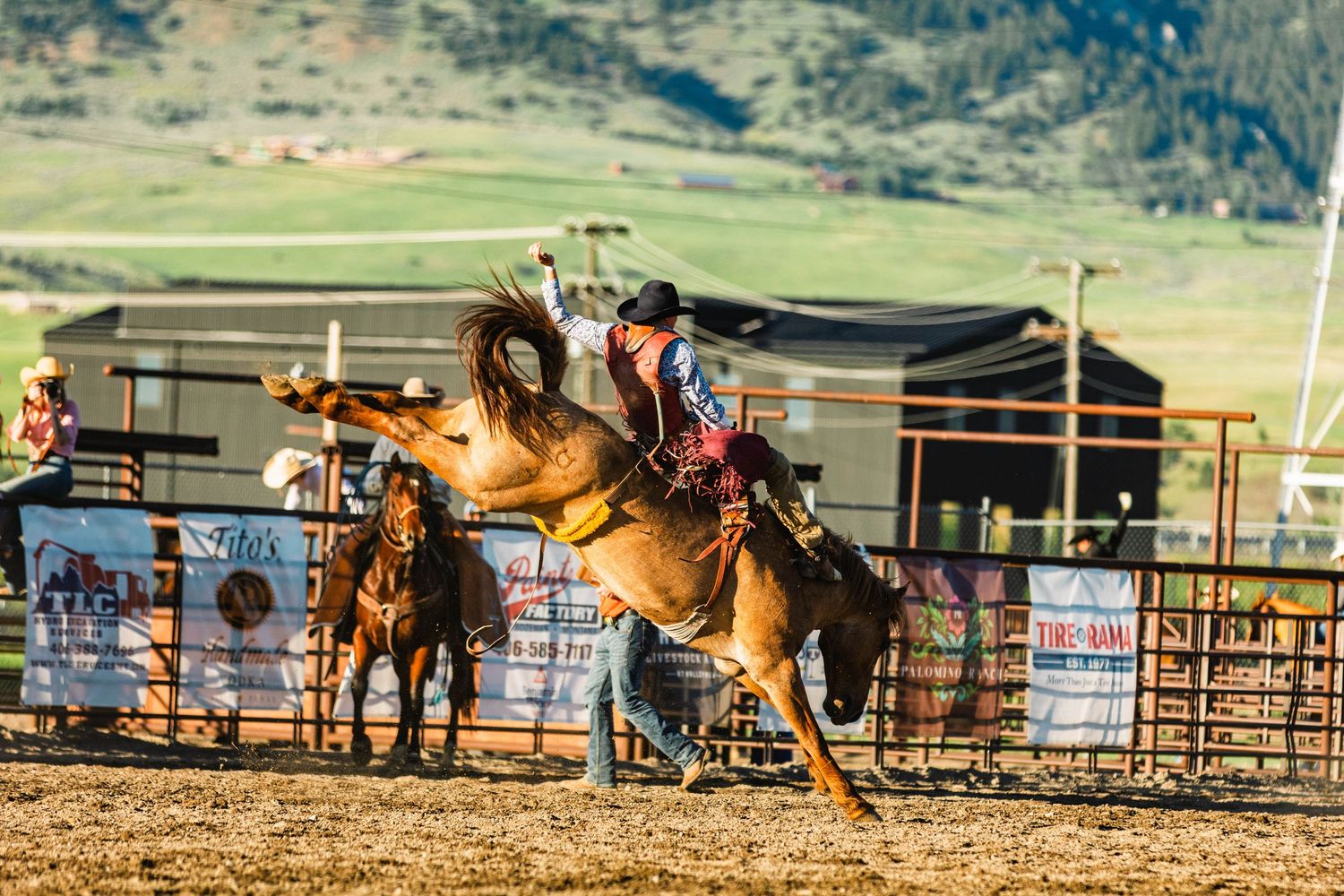 Cowboy rides a bucking bronco horse at an outdoor rodeo competition with mountains in the background.